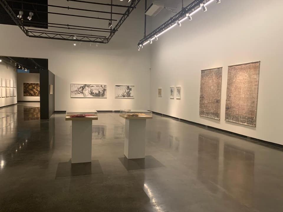 Art gallery with paintings on white walls, two pedestals in the center, and polished floor reflecting light.