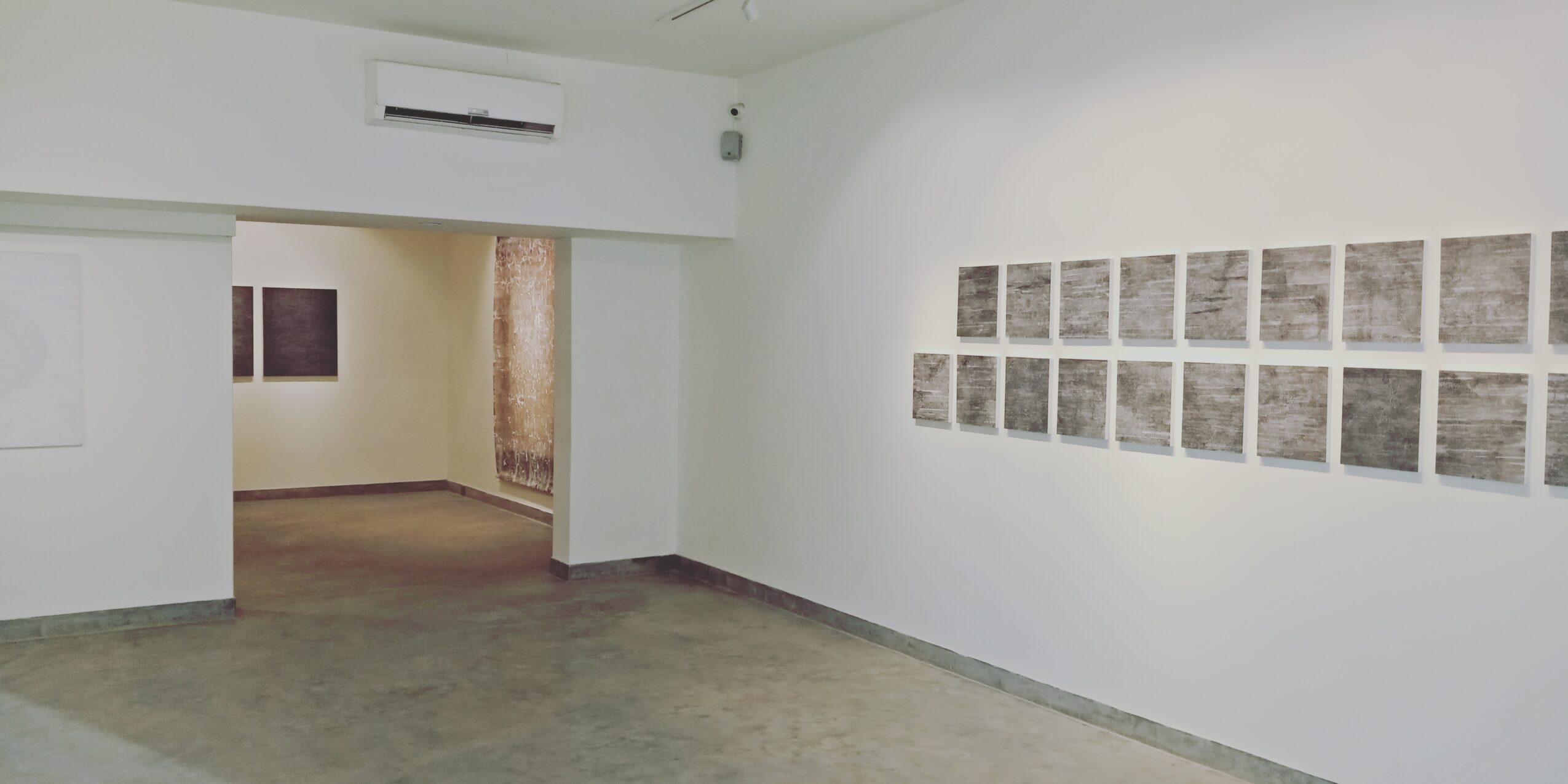 Empty art gallery with white walls, a series of small framed artworks, and a doorway leading to another room.