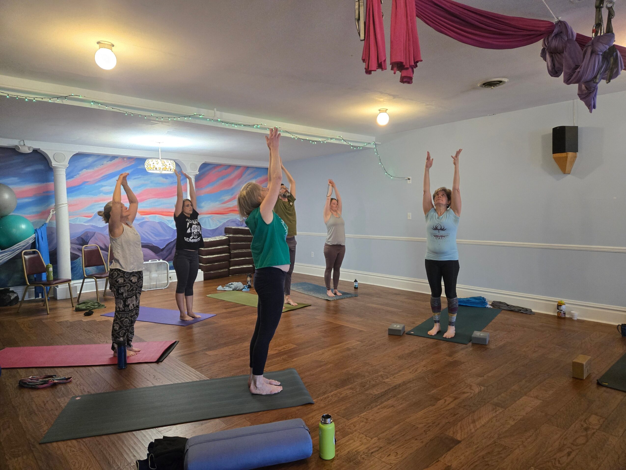 Group of six people practicing yoga with arms raised in a spacious room with wooden floor and colorful wall mural.