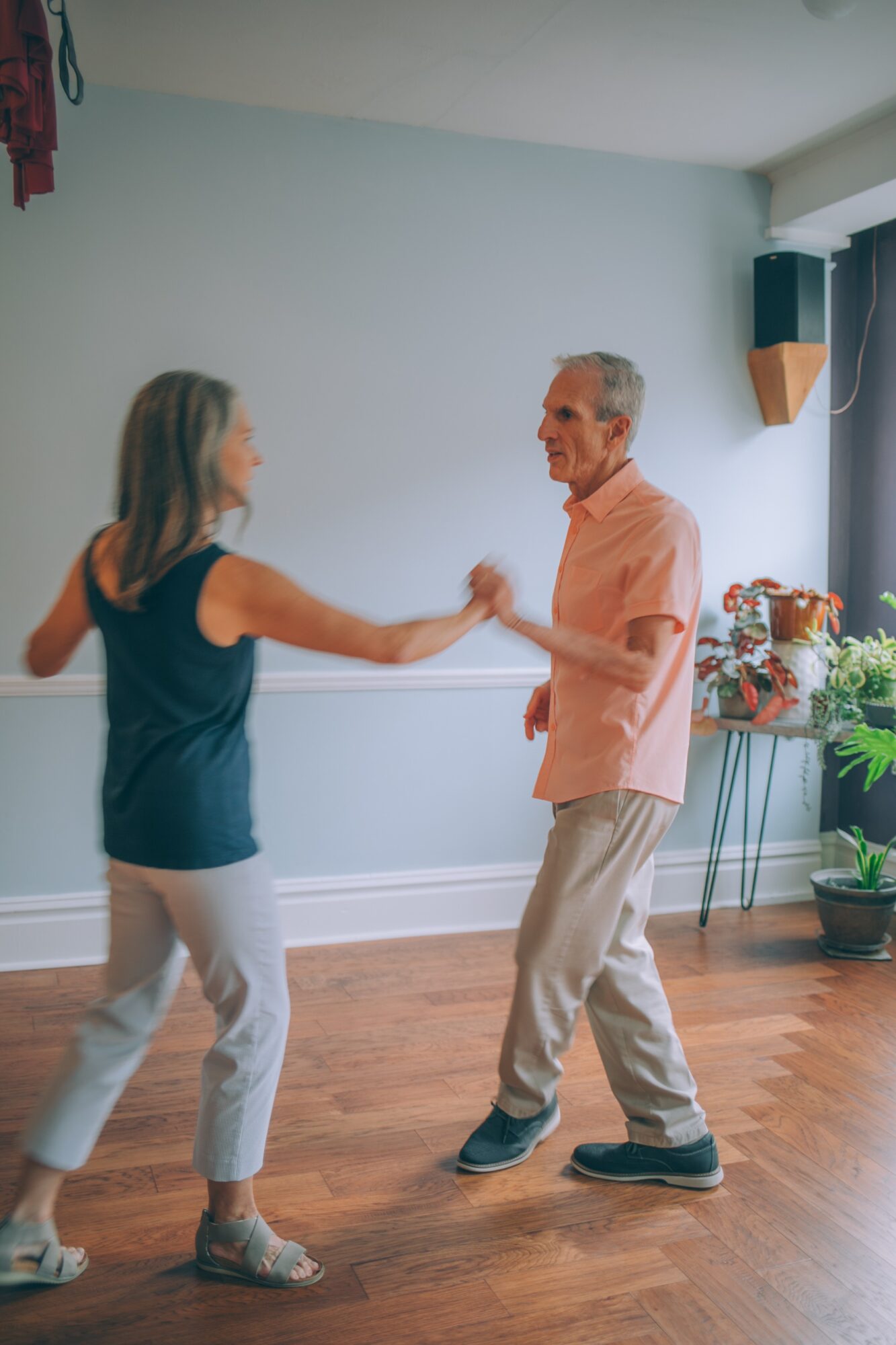 Two people dancing in a room with light blue walls, wooden floor, and a plant, holding hands and smiling.