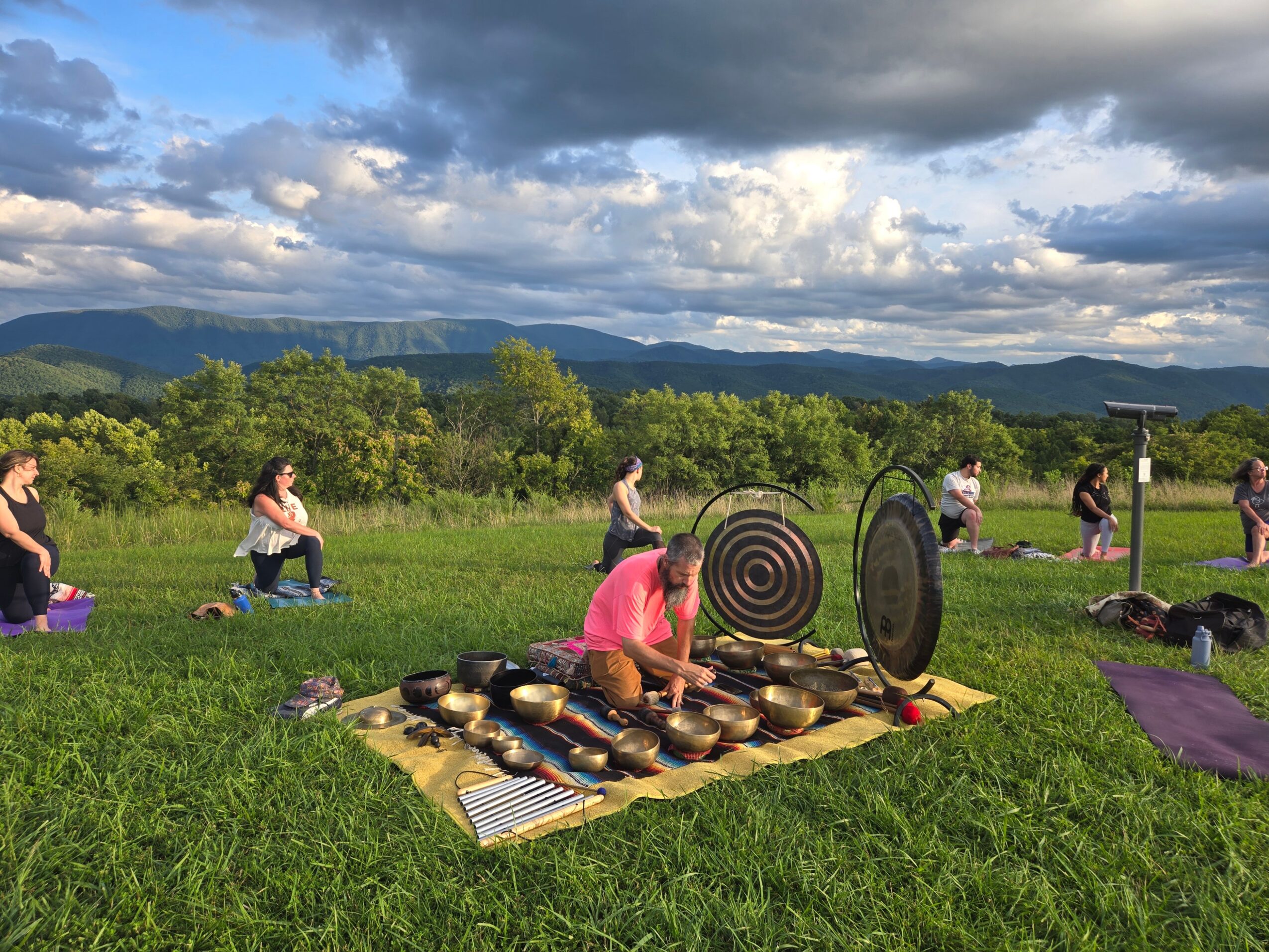 Group of people practicing yoga outdoors on a grassy field with mountains and cloudy sky in background.