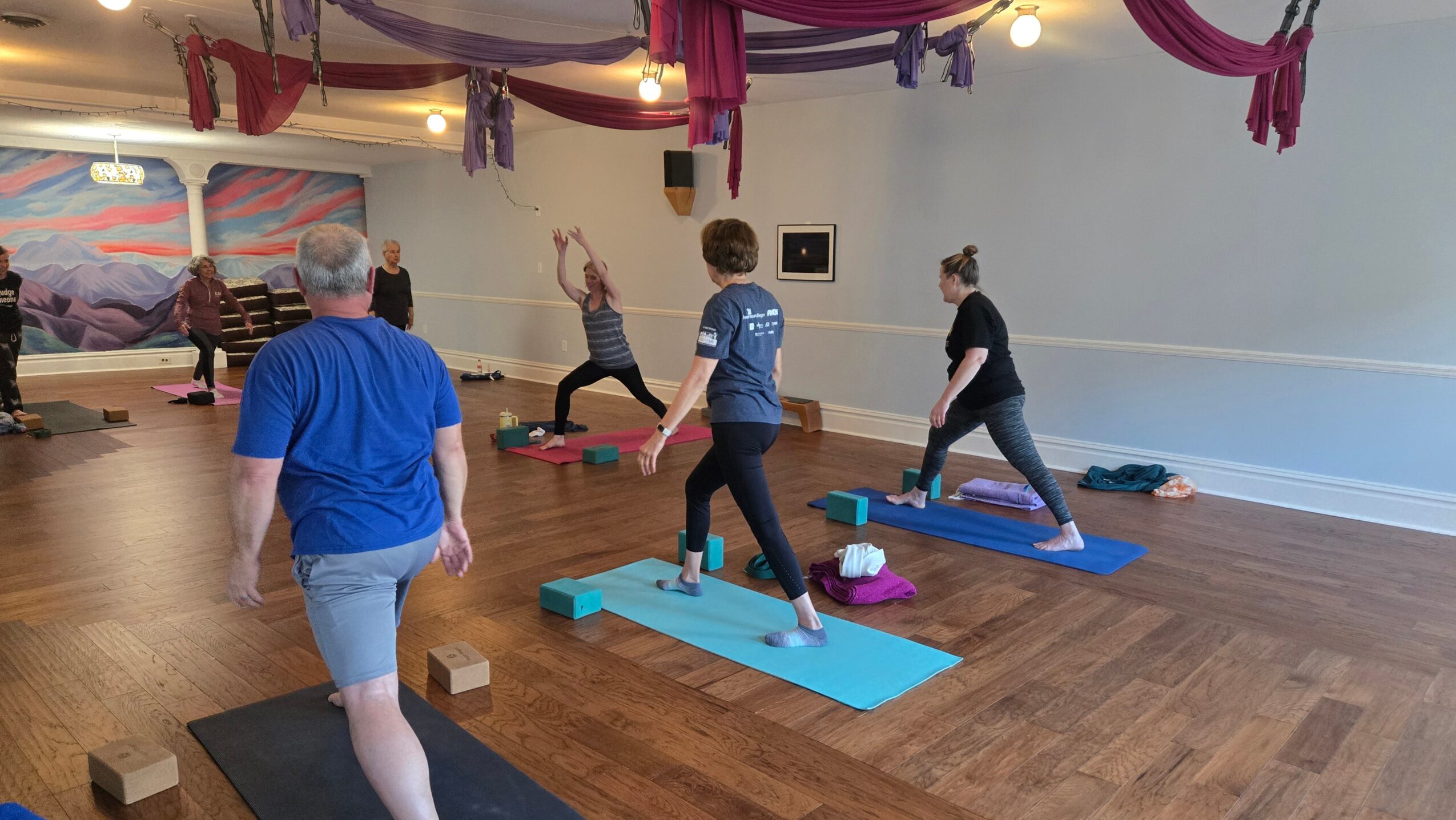 People practicing yoga on mats in a spacious room with wooden floors and colorful drapes hanging from the ceiling.
