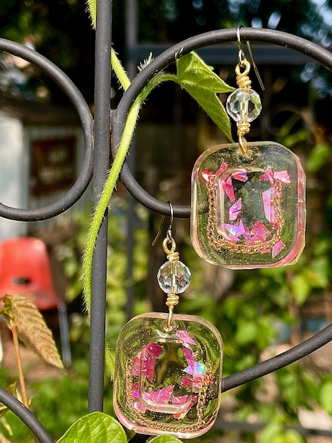 Two glass cube-shaped ornaments with pink designs hang from a metal plant stand, with a garden background.