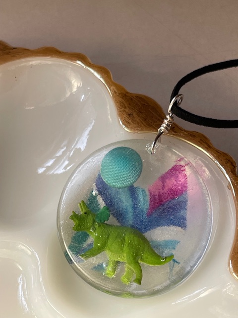 A small green toy dinosaur, a blue ball, and colorful foam pieces are inside a clear container of water.