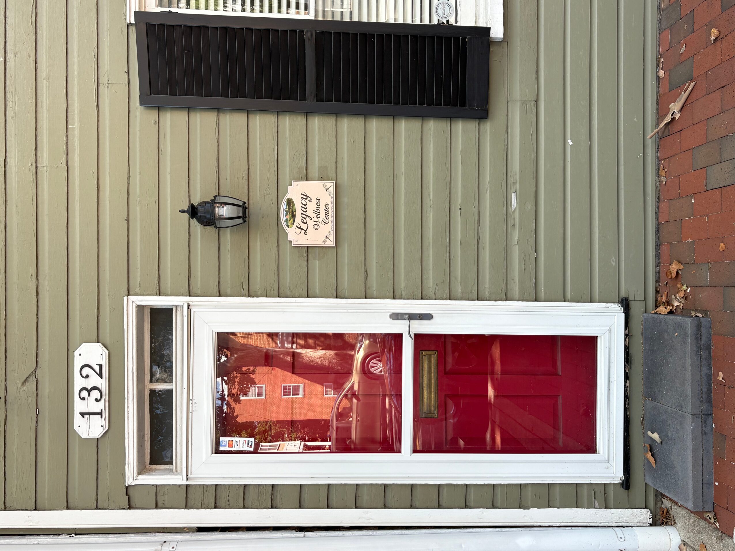 Front door with a large glass window, white frame, and a red curtain inside, set in a green wall with a brick pathway.