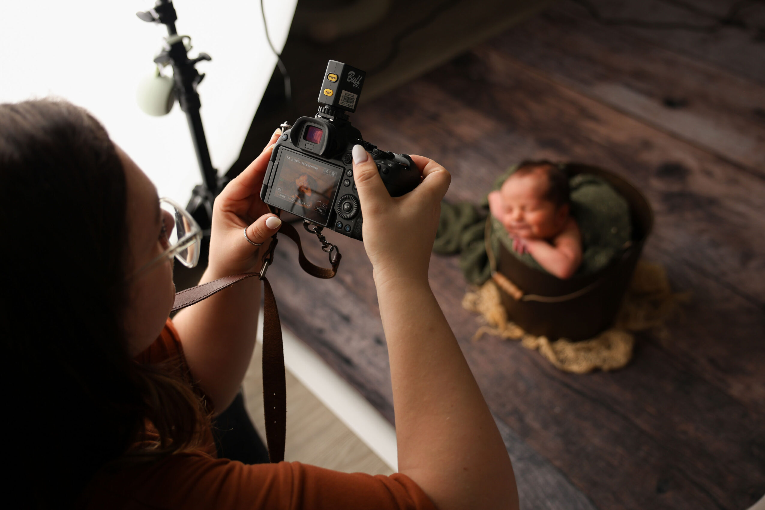 Person taking a photo of a baby sitting in a basket on a wooden floor, with photography equipment visible.