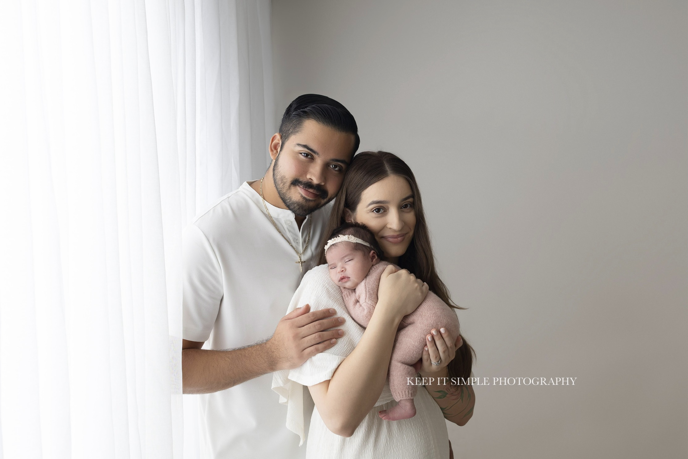 Family of three with a newborn, woman holding baby, man and woman smiling indoors near window, light-colored wall background.