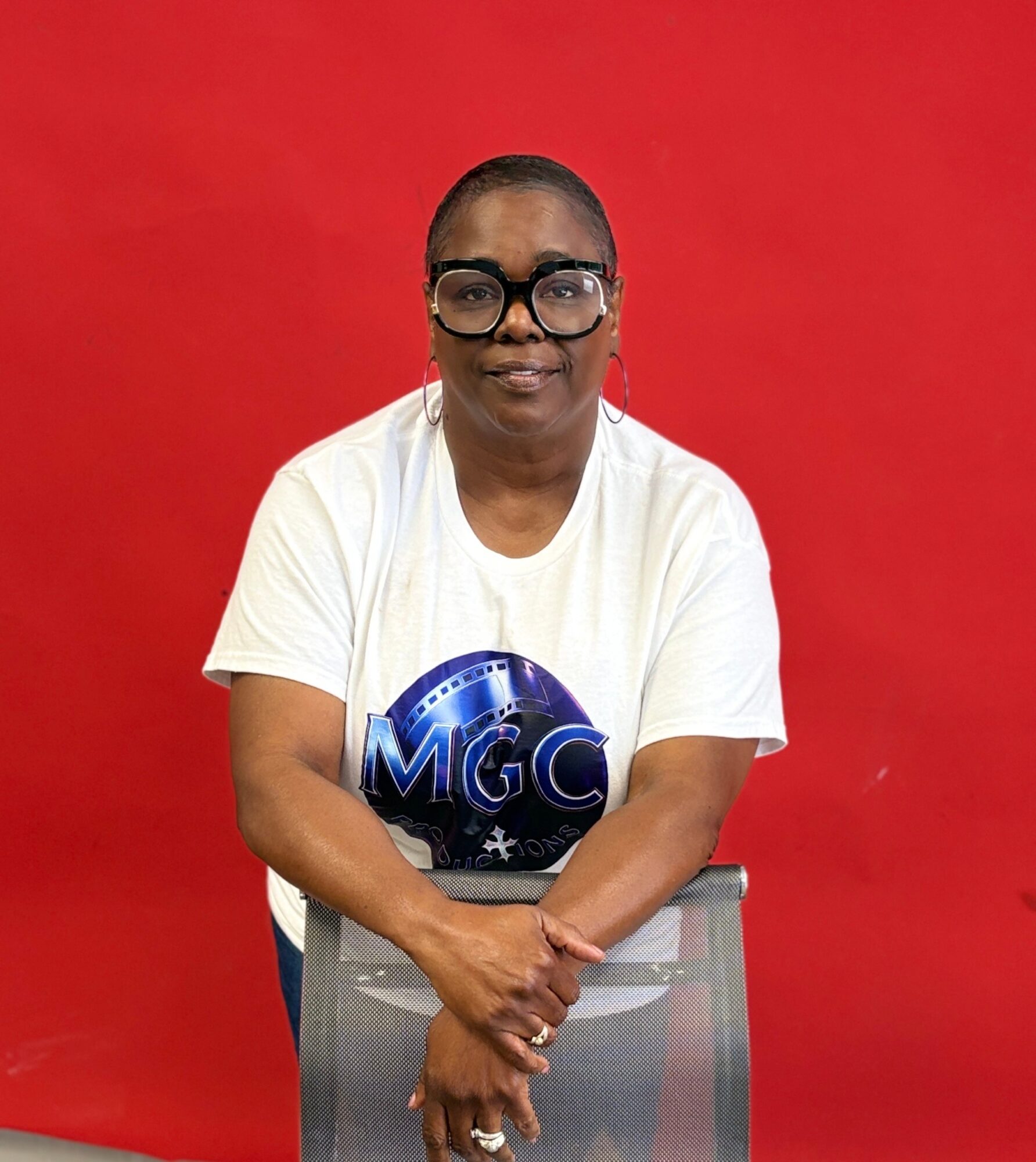 Woman with glasses and earrings sitting in front of a red background, wearing a white T-shirt with a blue logo, arms crossed.