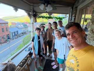 Group of people, including children and adults, gathered on a porch with a view of a street and buildings outside.