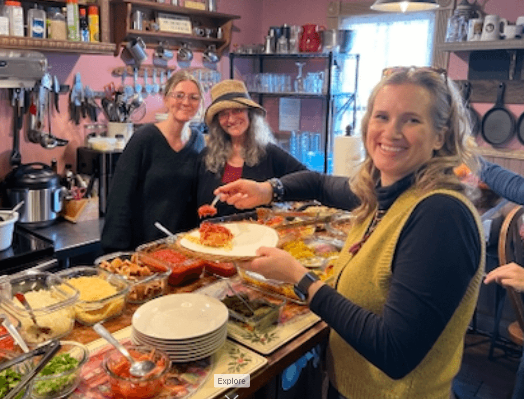 Three women smiling in a kitchen, one serving food onto a plate, surrounded by dishes and kitchen utensils.