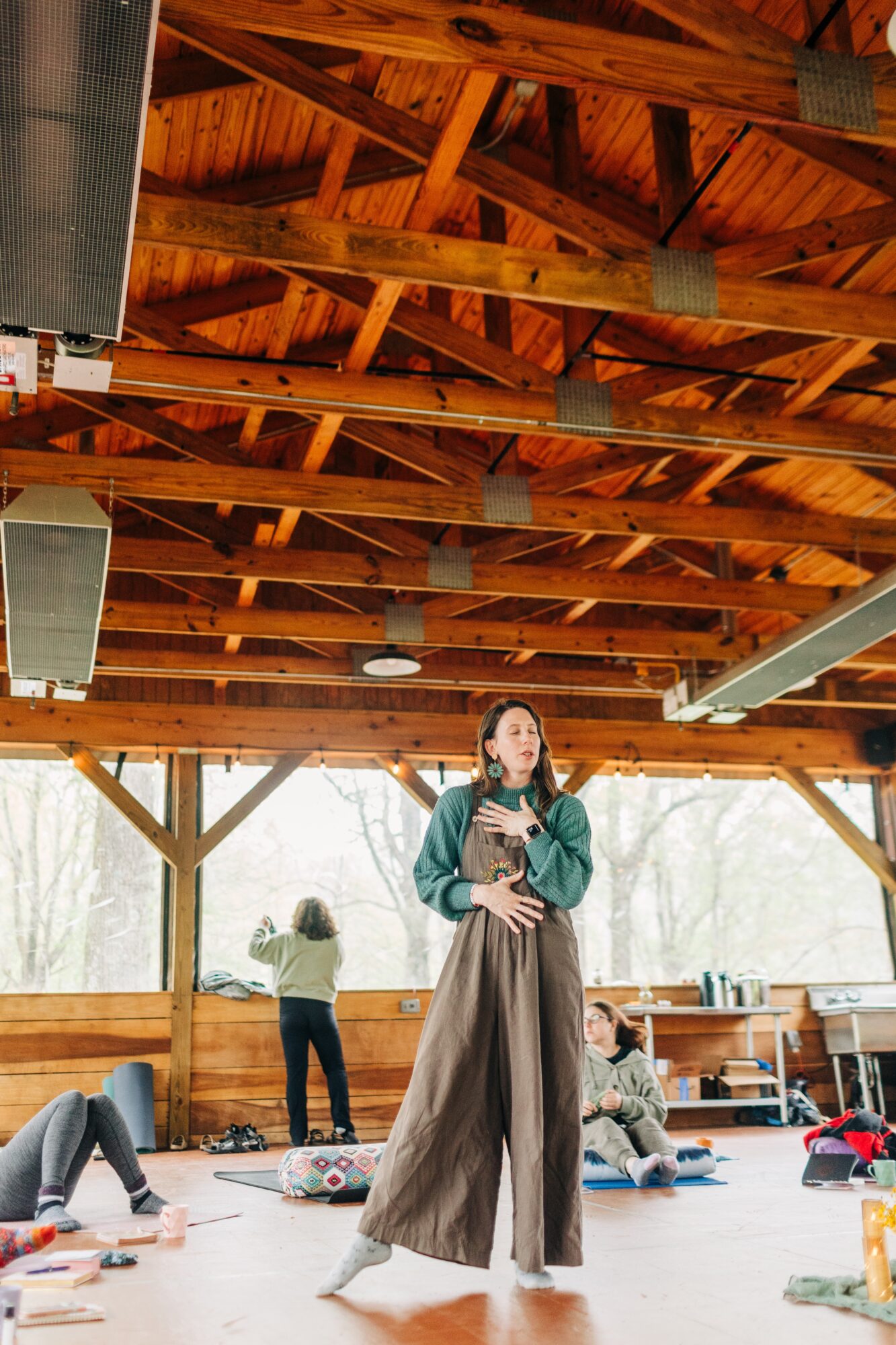 Woman standing in a spacious wooden indoor area with large windows, people sitting and standing around, some with yoga mats.