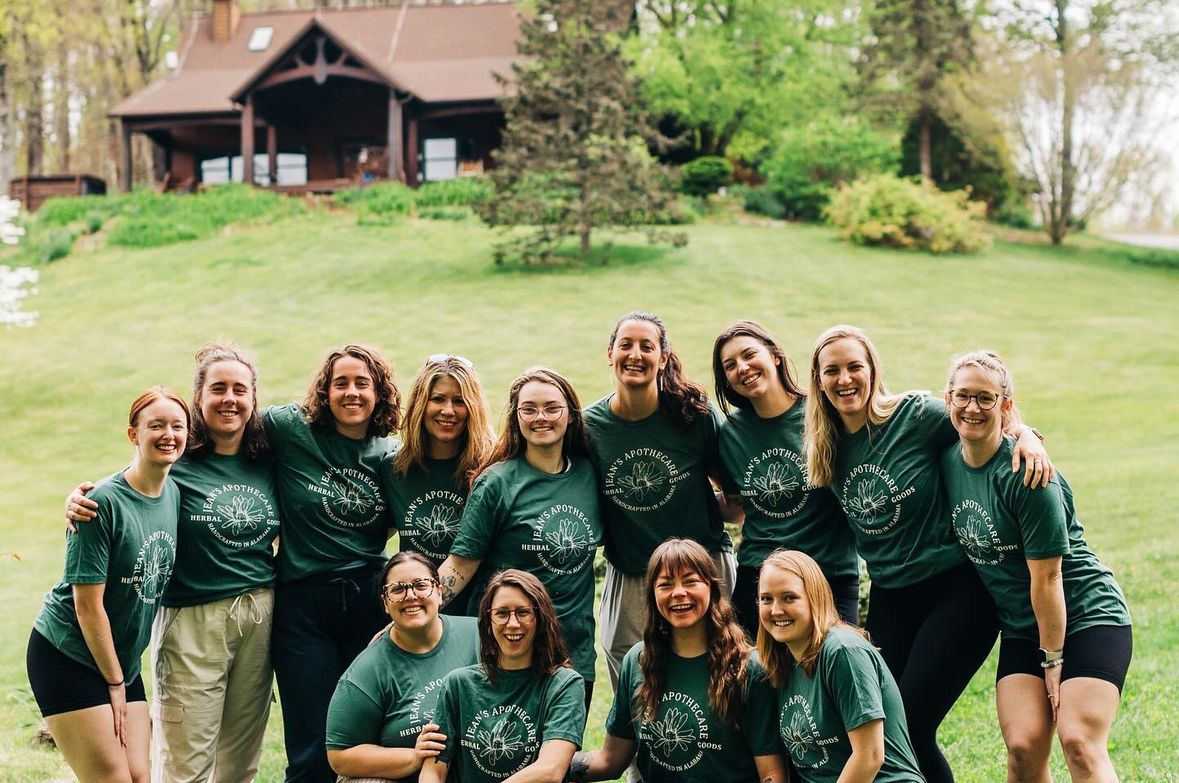 Group of ten people outdoors on a grassy hill, smiling, wearing matching green shirts, with trees and a house in the background.