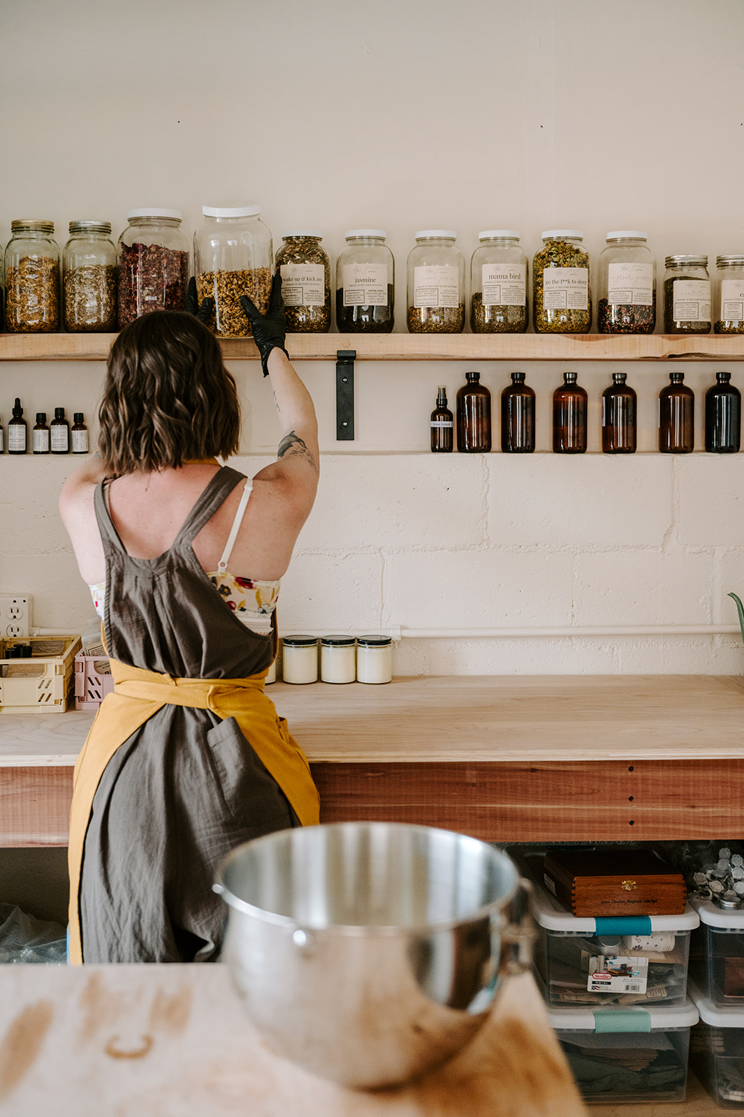 Person with dark hair wearing an apron reaches for jars on a shelf in a kitchen. A metal bowl is on the counter.