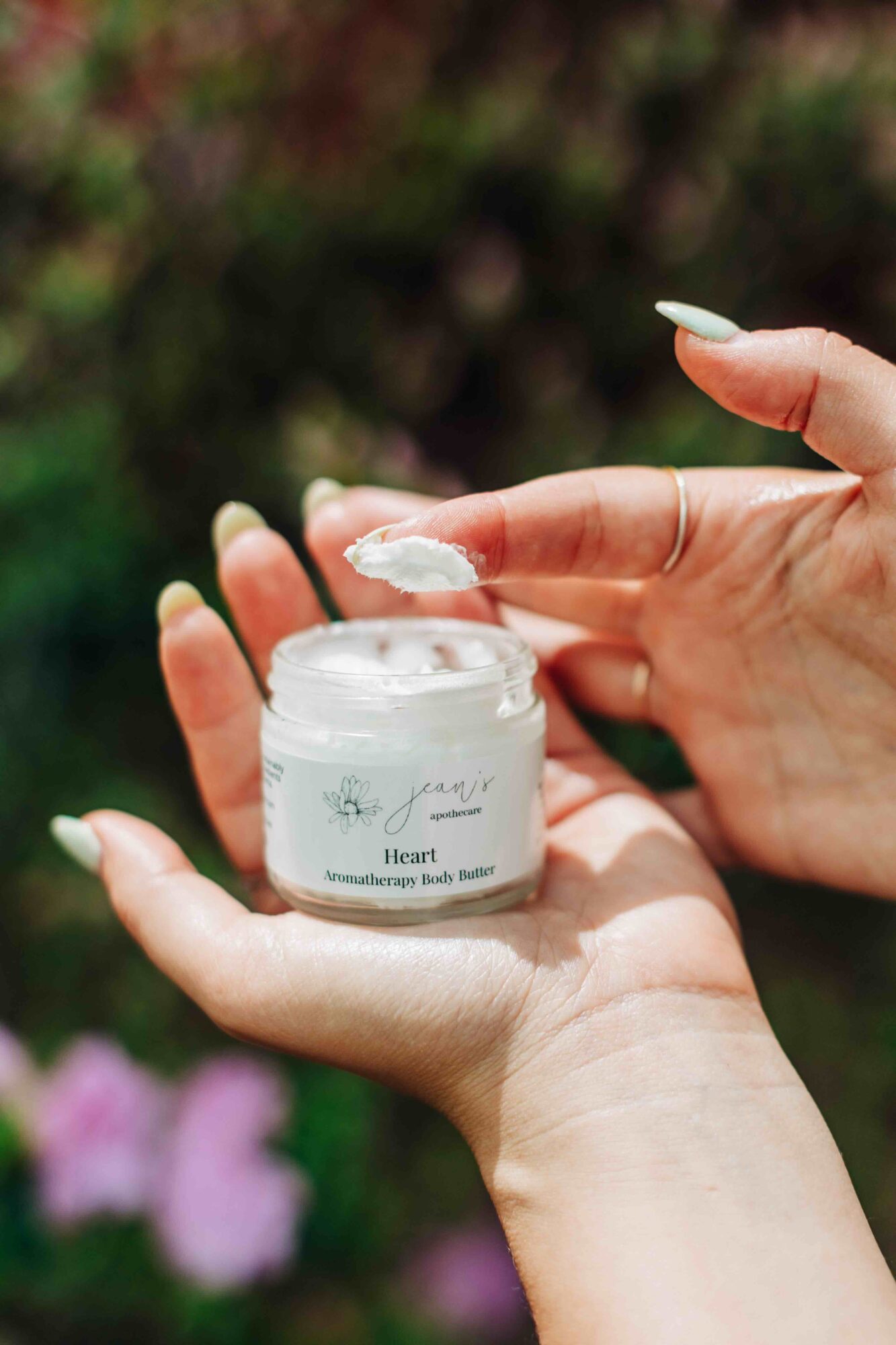 Person applying white cream from jar with a garden background, pink flowers visible, hand holding jar, other hand spreading cream.