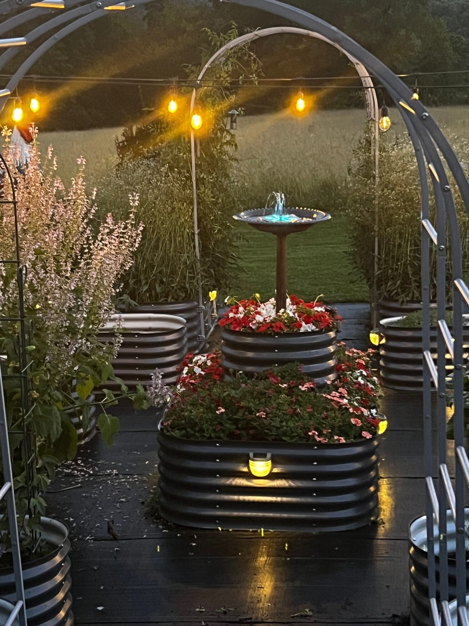 Decorative fountain with flowers and candles, surrounded by plants and outdoor lighting, during evening or dusk.