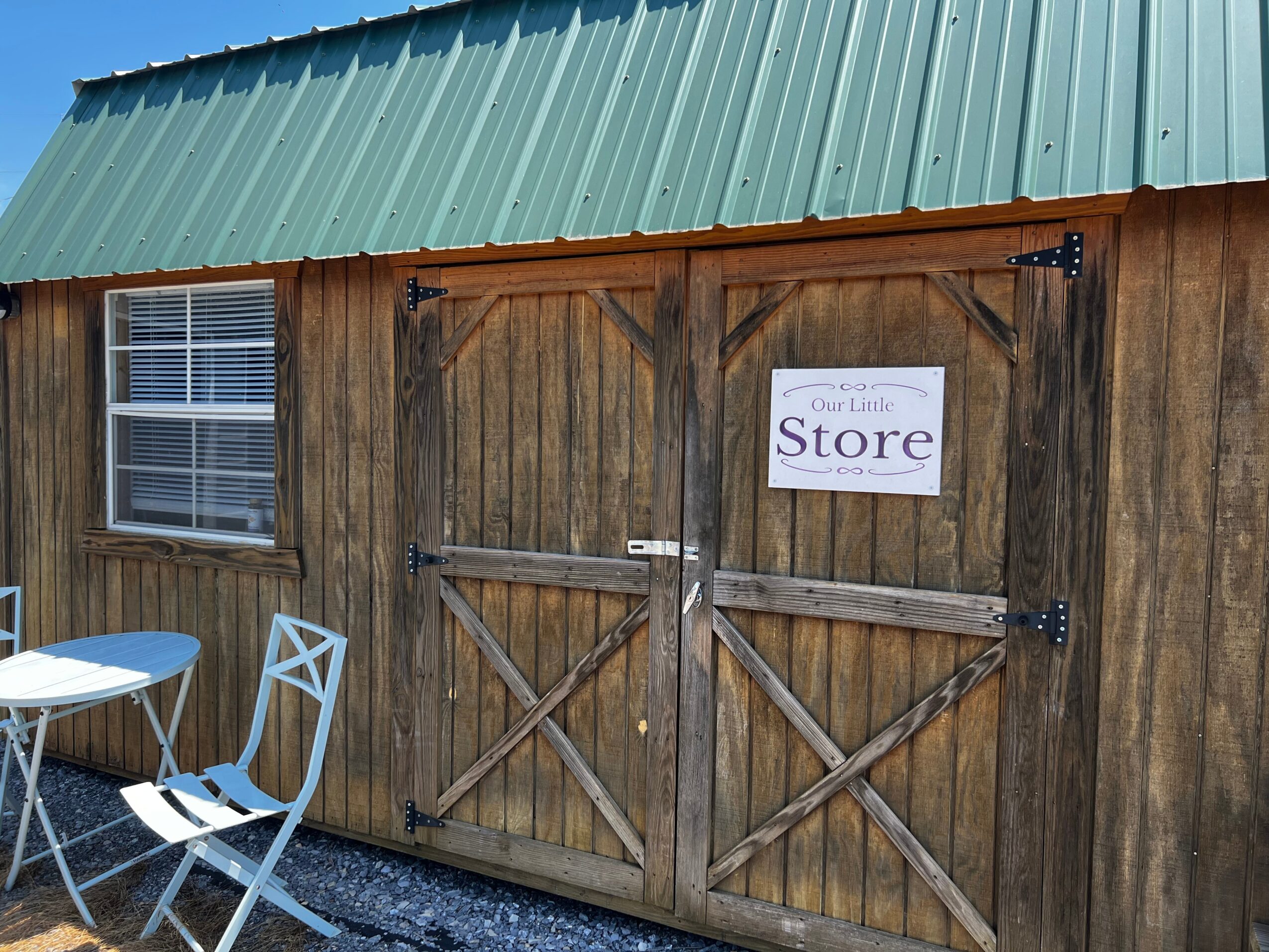 Wooden building with a green metal roof, window, and a double door with a store sign, outdoor table and chairs.
