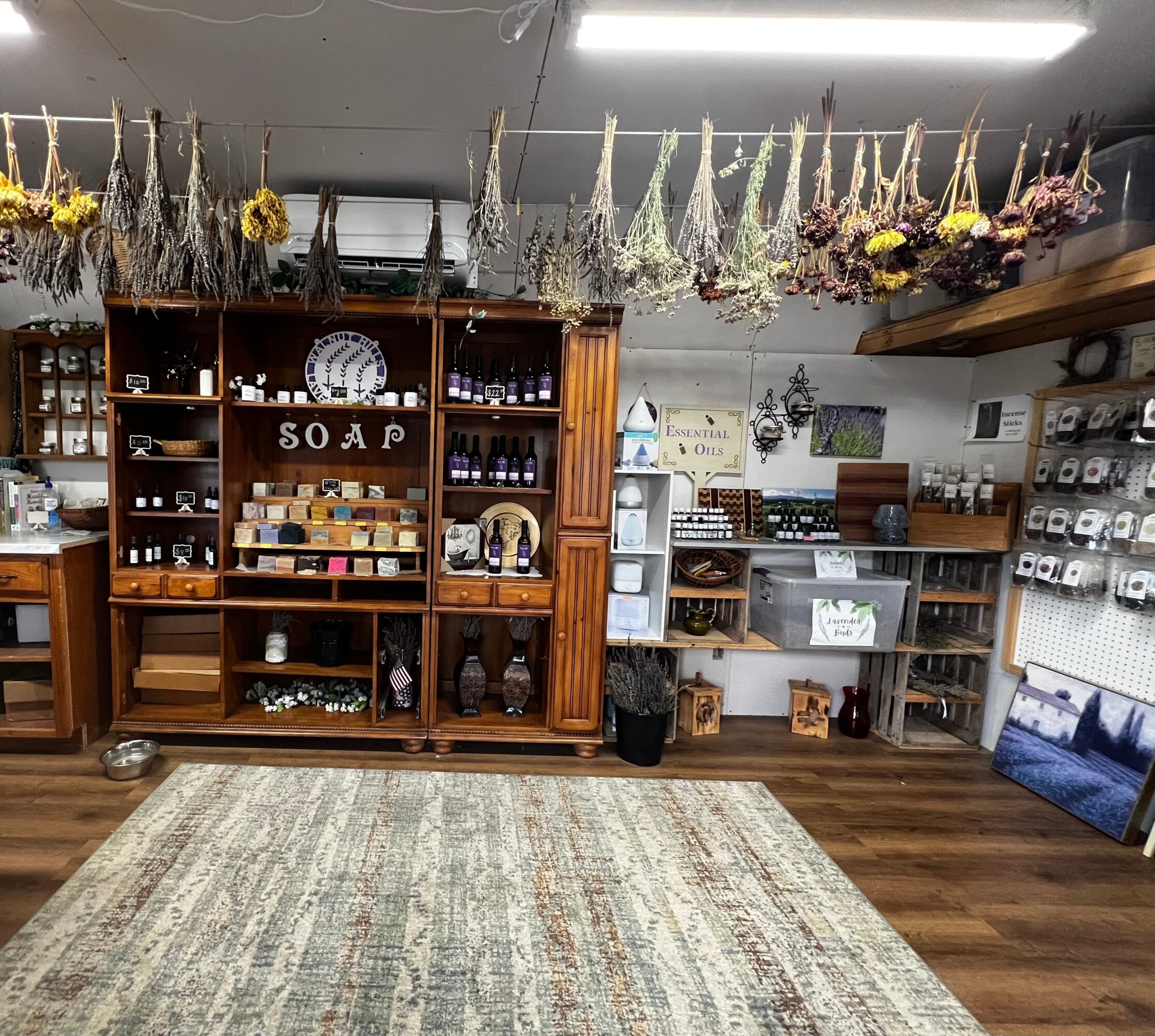 Interior of a store with wooden shelves displaying various items, hanging dried flowers, and a large rug on the floor.