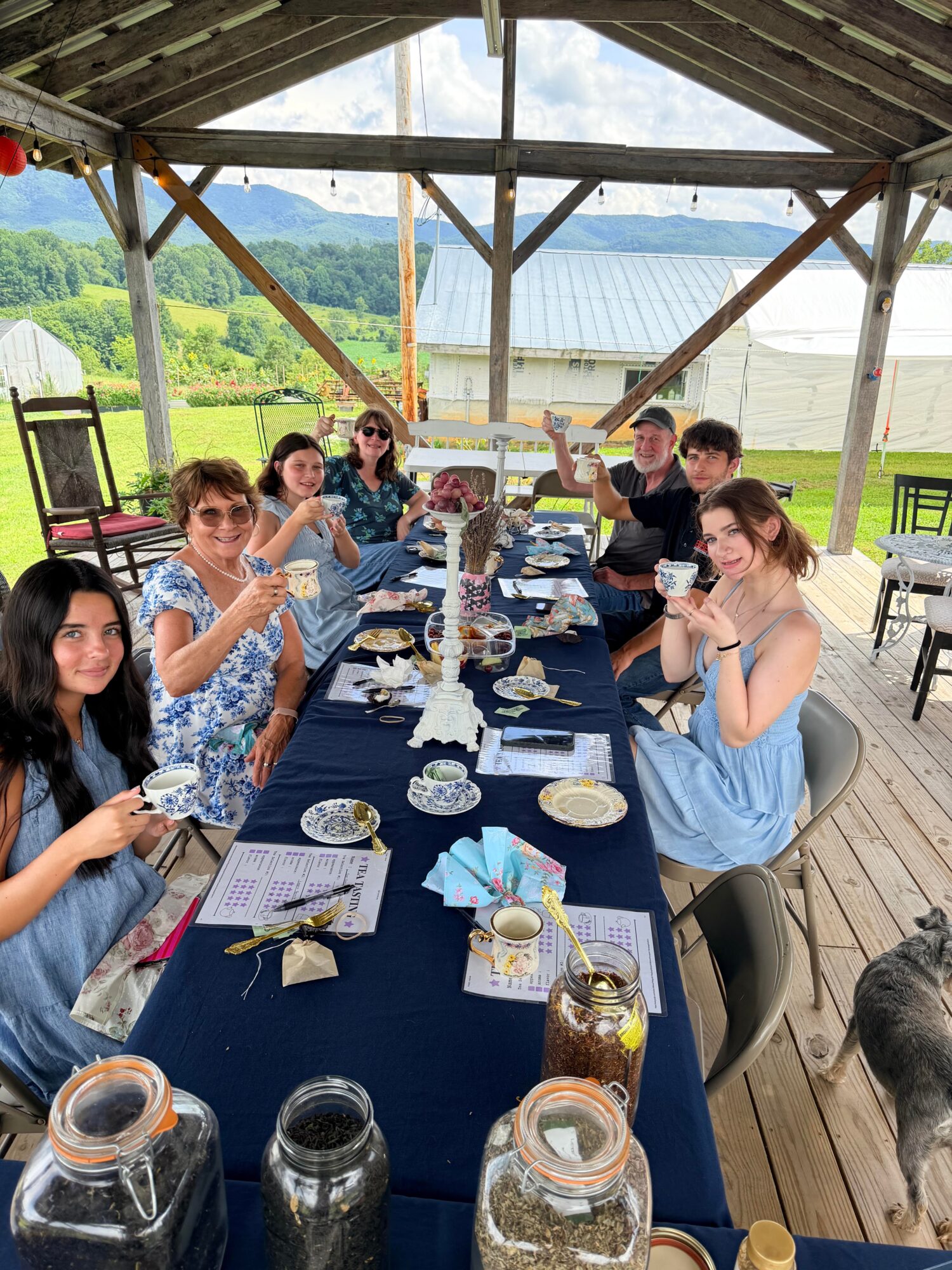 Group of people sitting at a long table outdoors under a wooden pavilion, enjoying a meal with greenery in the background.