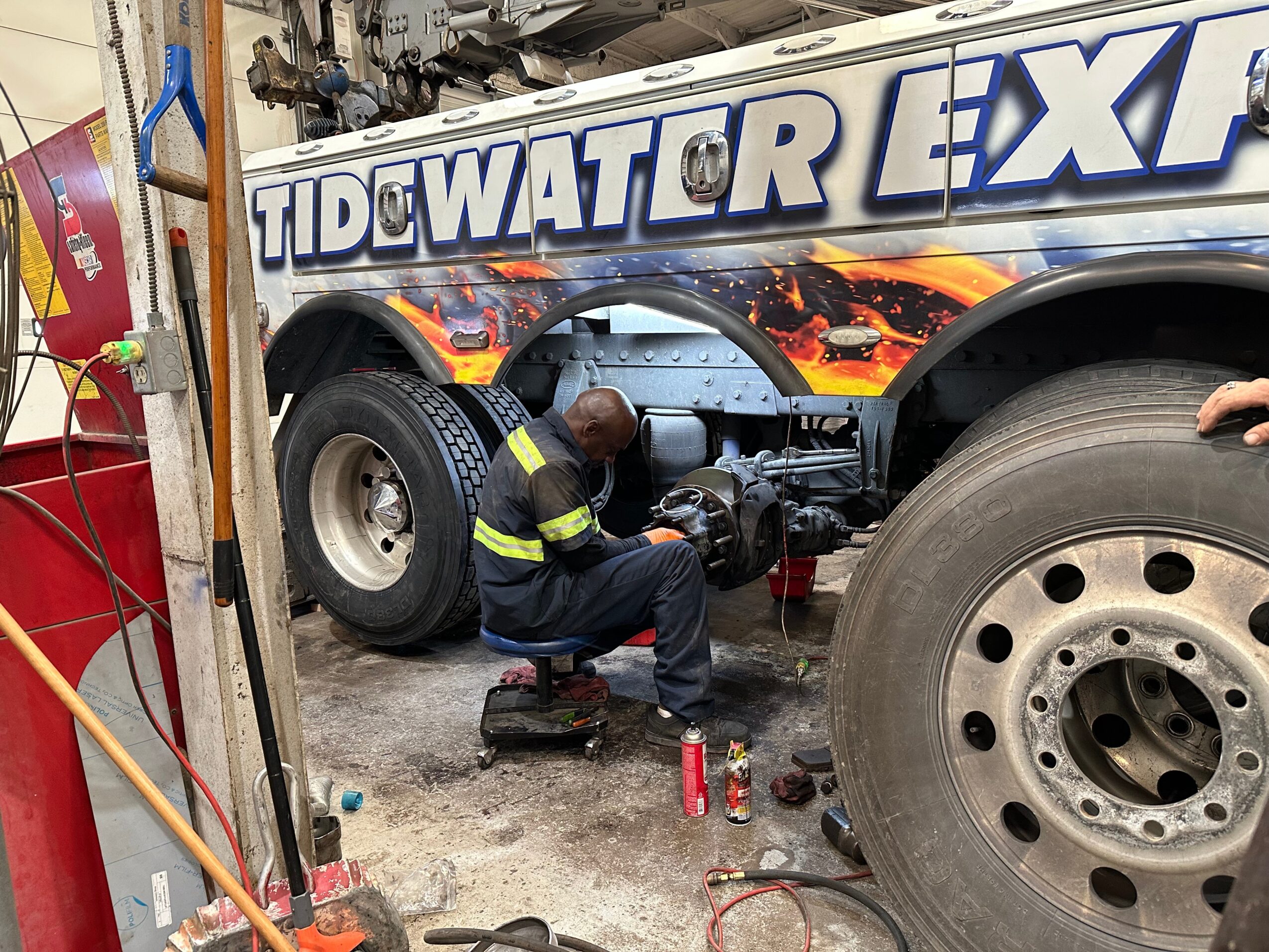 Worker in uniform repairing a large fire truck with colorful graphics and text, tools and equipment on the ground.