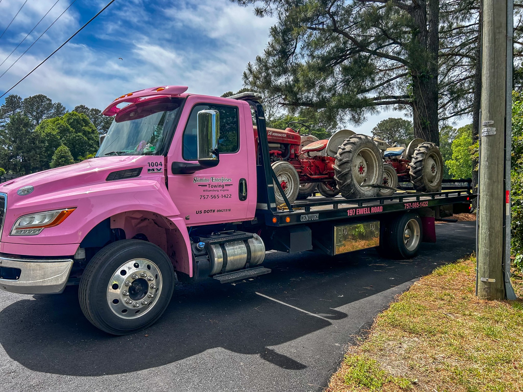 Pink tow truck carrying a red tractor with large tires parked on a street with trees and a utility pole nearby.