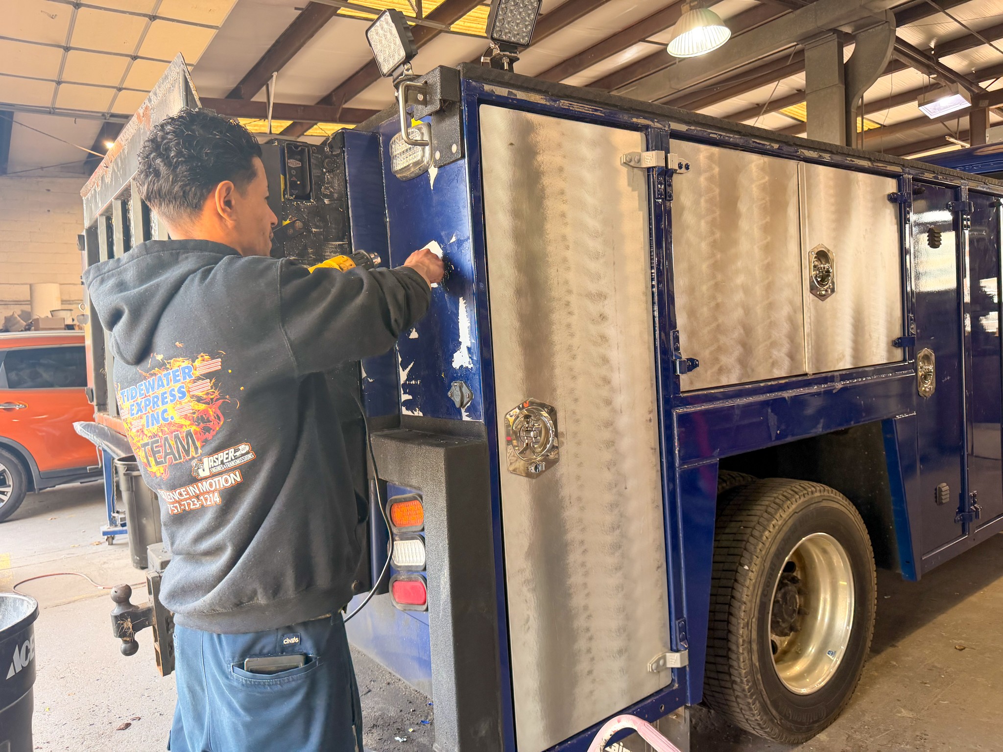 Person working on a large blue utility truck inside a garage, with a vehicle visible in the background.