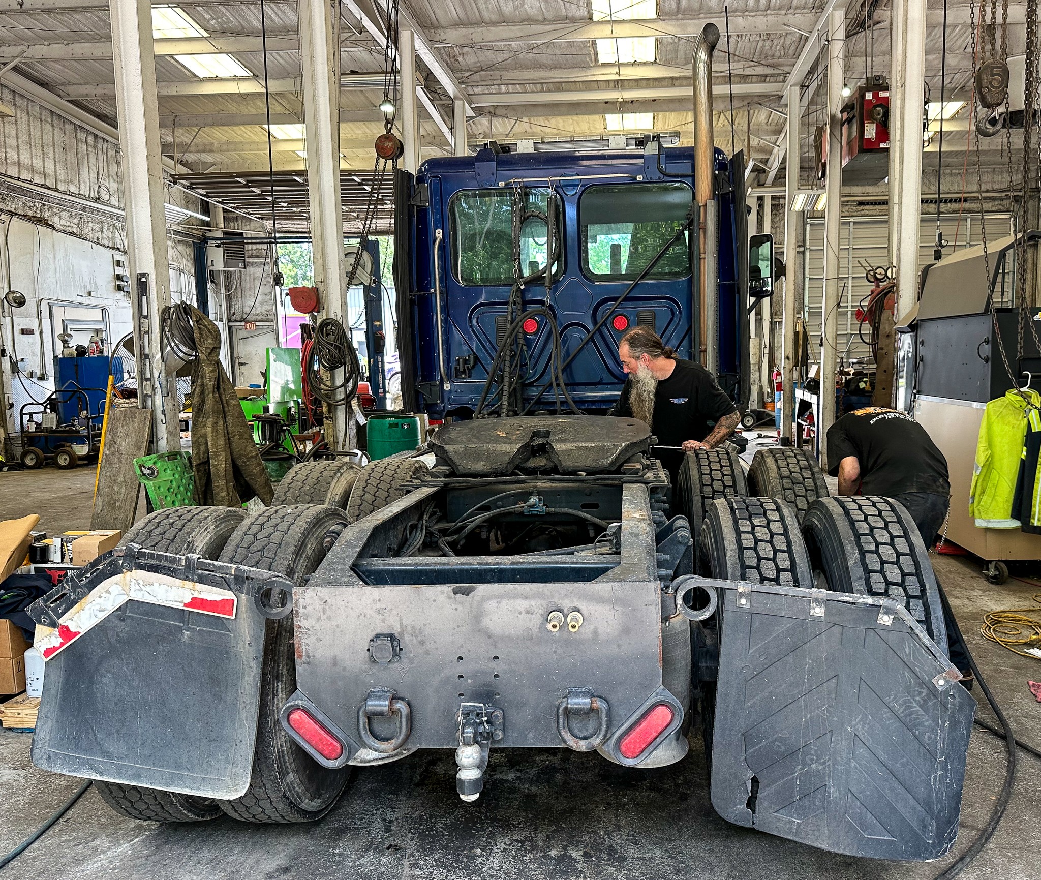 Inside a workshop, a large blue truck is being serviced with two workers attending to it, surrounded by tools and equipment.