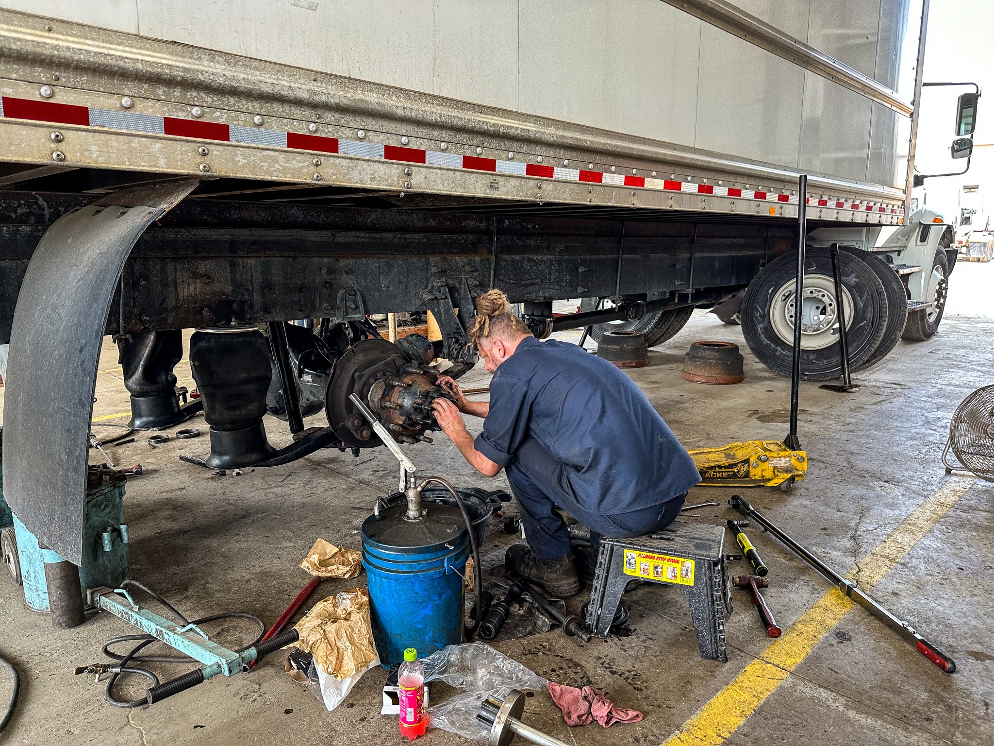 A person working underneath a large truck, using tools near the truck's wheel, with equipment and supplies around.