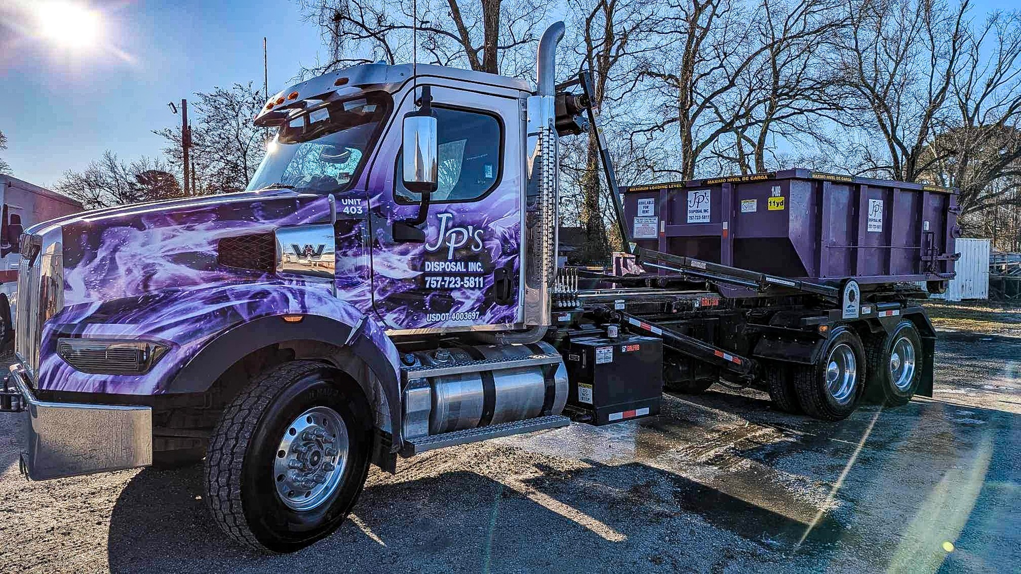 Semi-truck with purple and black design parked in a lot, with trees and a clear sky in the background.