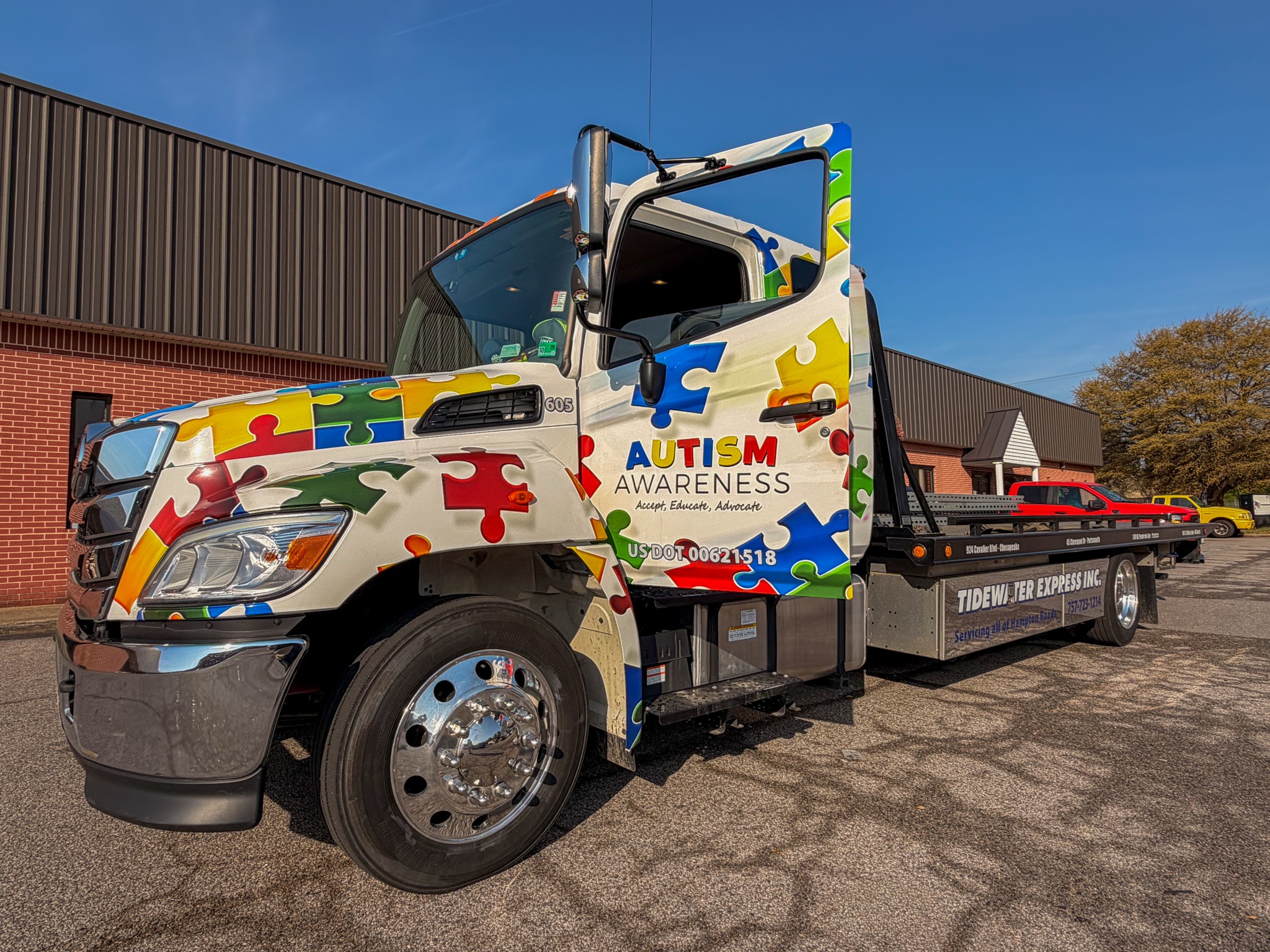 Flatbed truck with colorful puzzle piece design and autism awareness message parked outdoors, buildings and trees in background.
