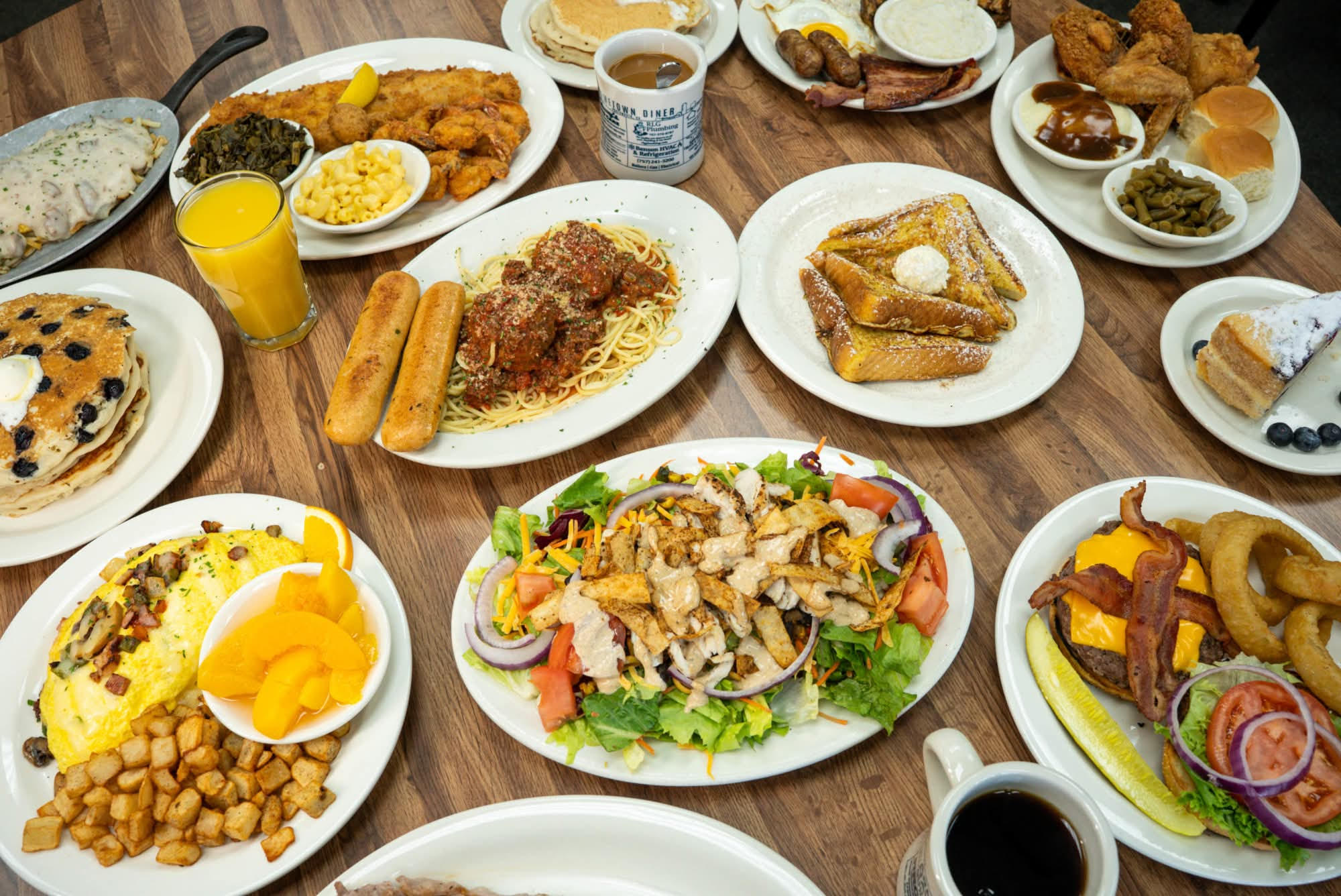 Various dishes including fried chicken, bread, salad, and beverages on a wooden table, with a glass of orange juice and coffee.
