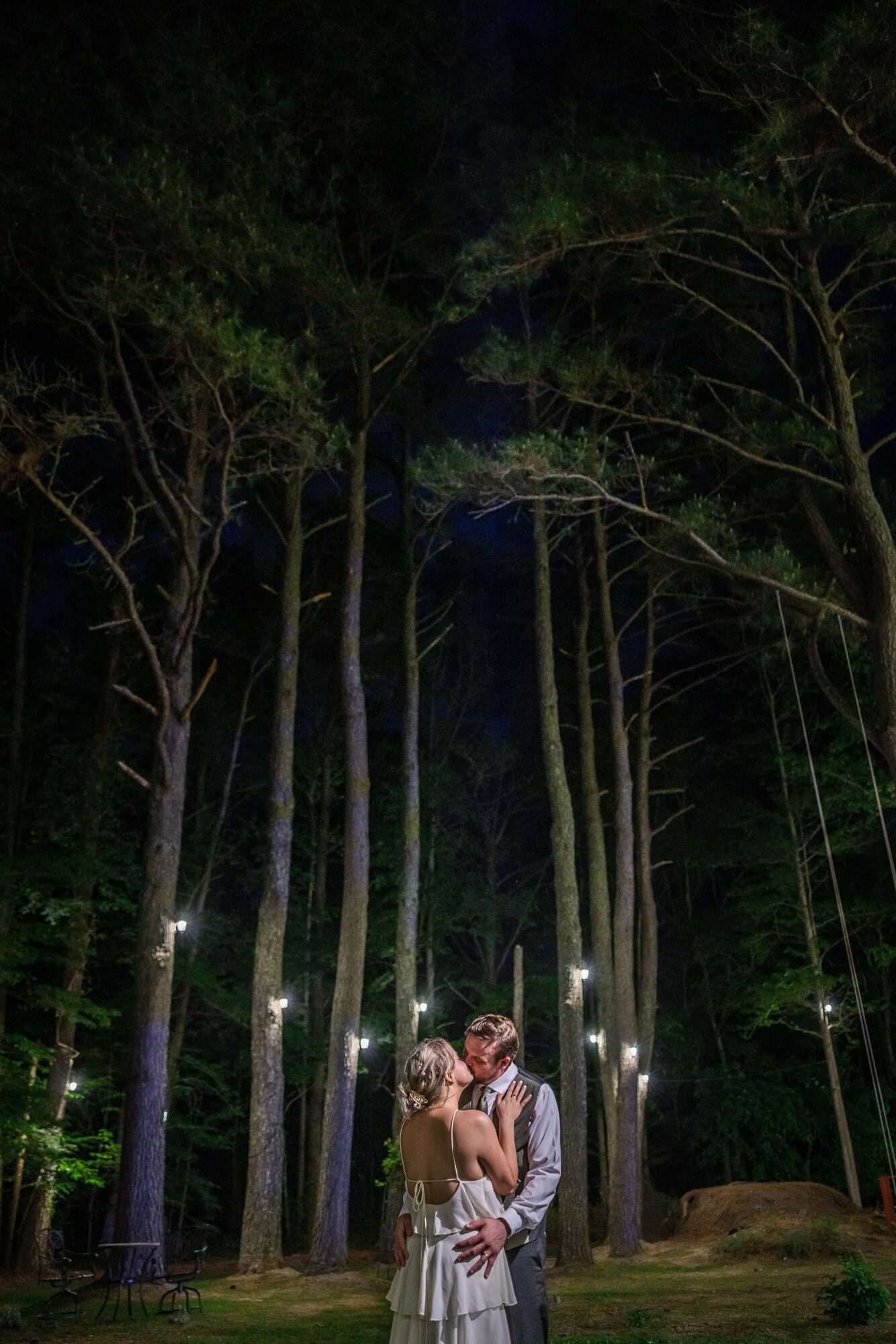Couple embracing in a forest at night with tall trees and small lights around them.