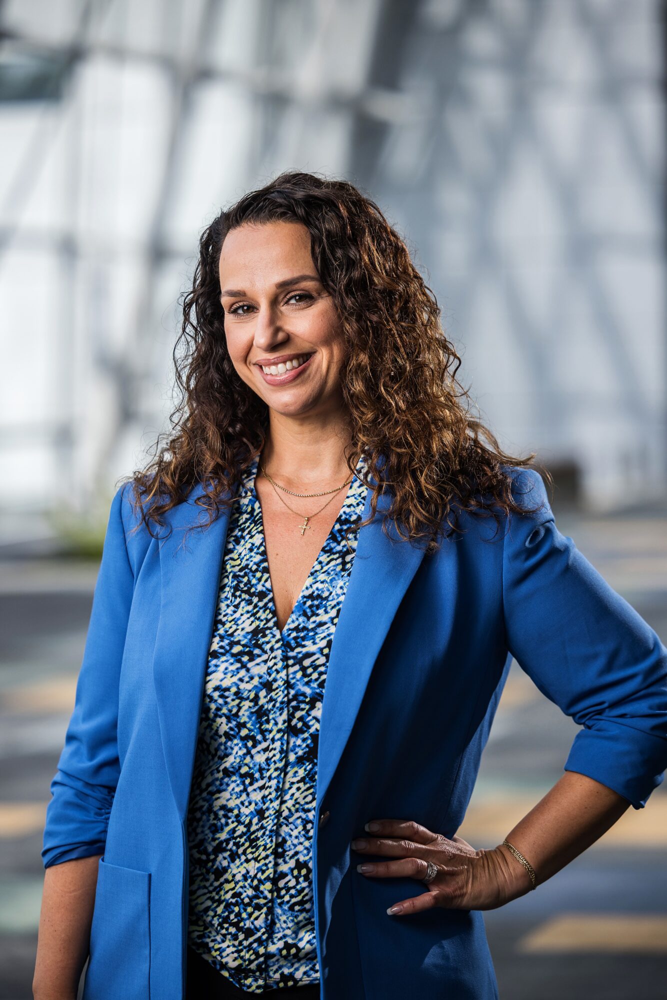 Woman with curly hair smiling, wearing a blue blazer and patterned blouse, standing indoors with a modern glass background.
