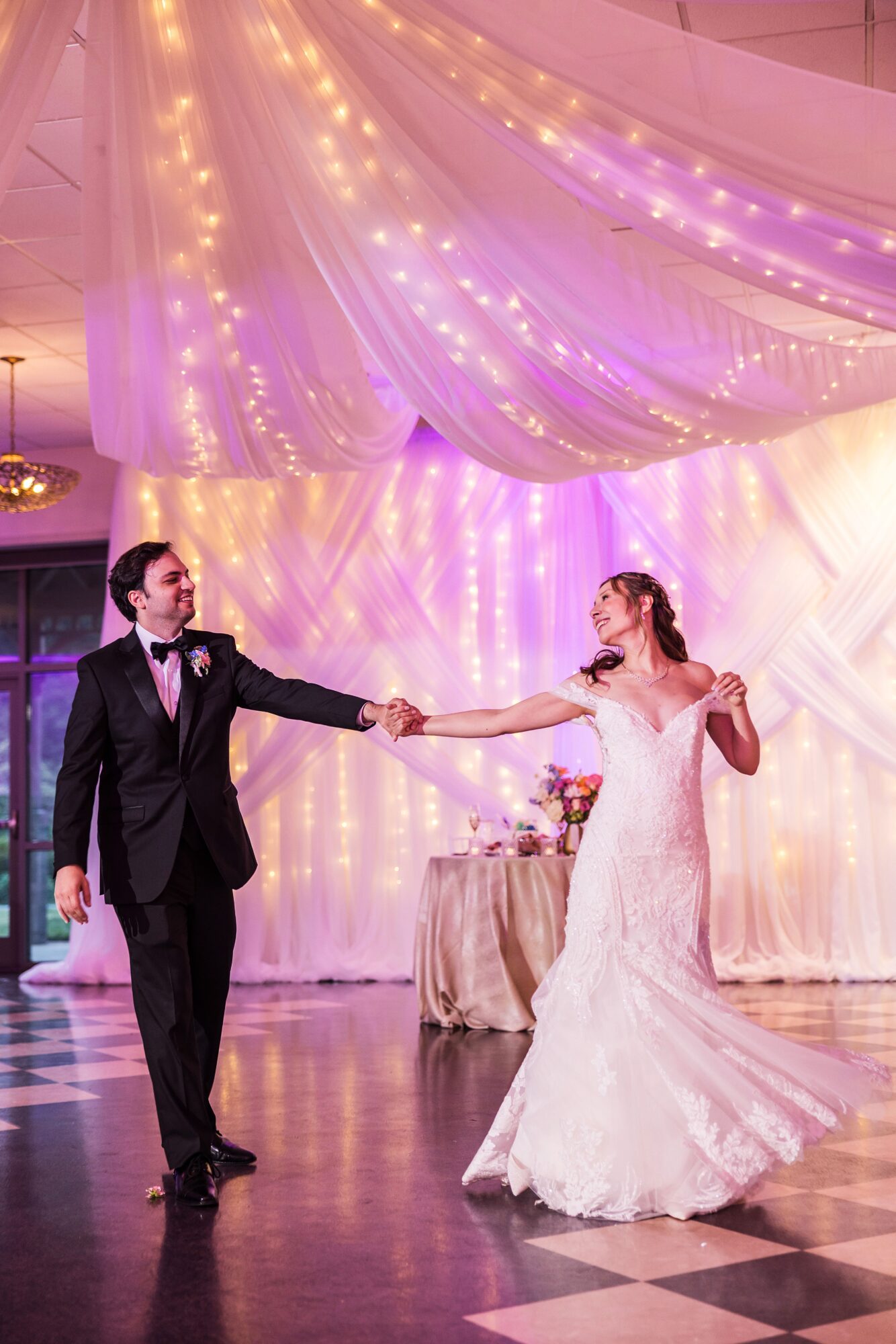A bride and groom dance at their wedding reception, holding hands, with a decorated backdrop and table in the background.