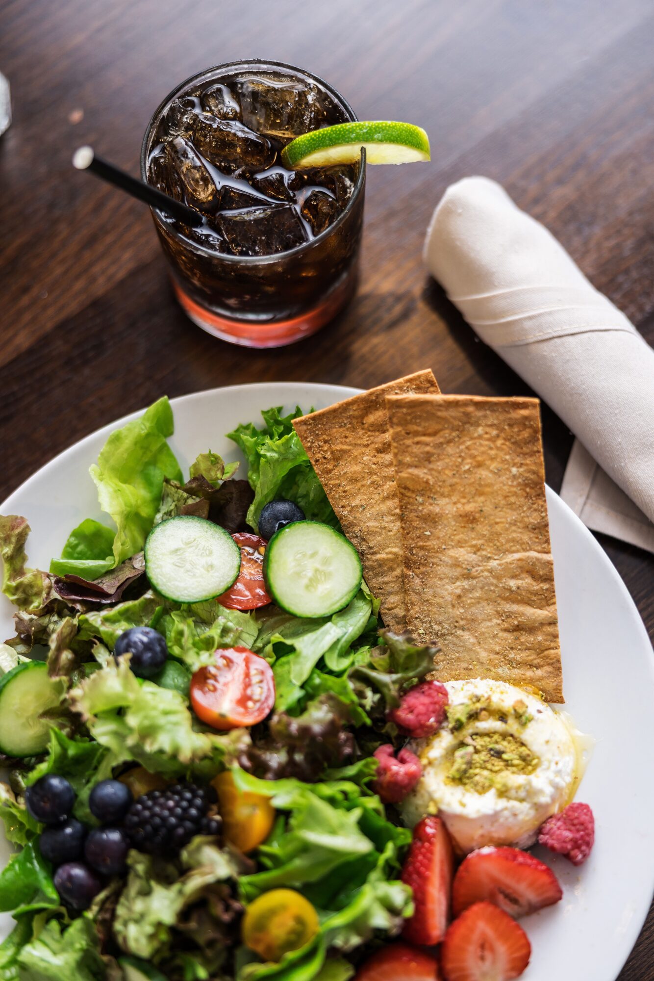 Glass of soda with lime wedge, plate of salad with cucumbers, strawberries, blueberries, and crackers, on a wooden table.
