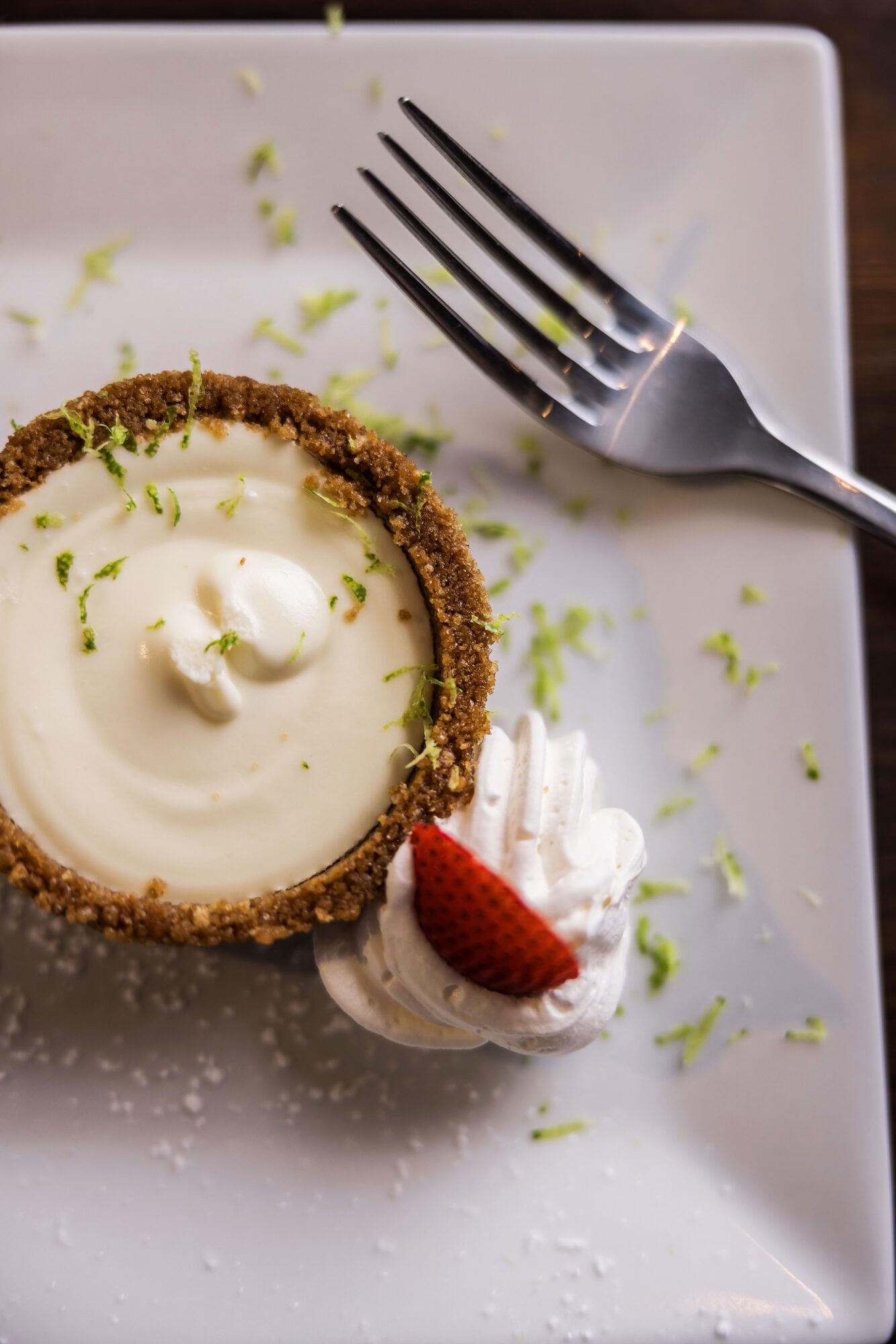 Dessert with a crusted tart shell, creamy filling, garnished with a small strawberry and whipped cream, on a white plate.