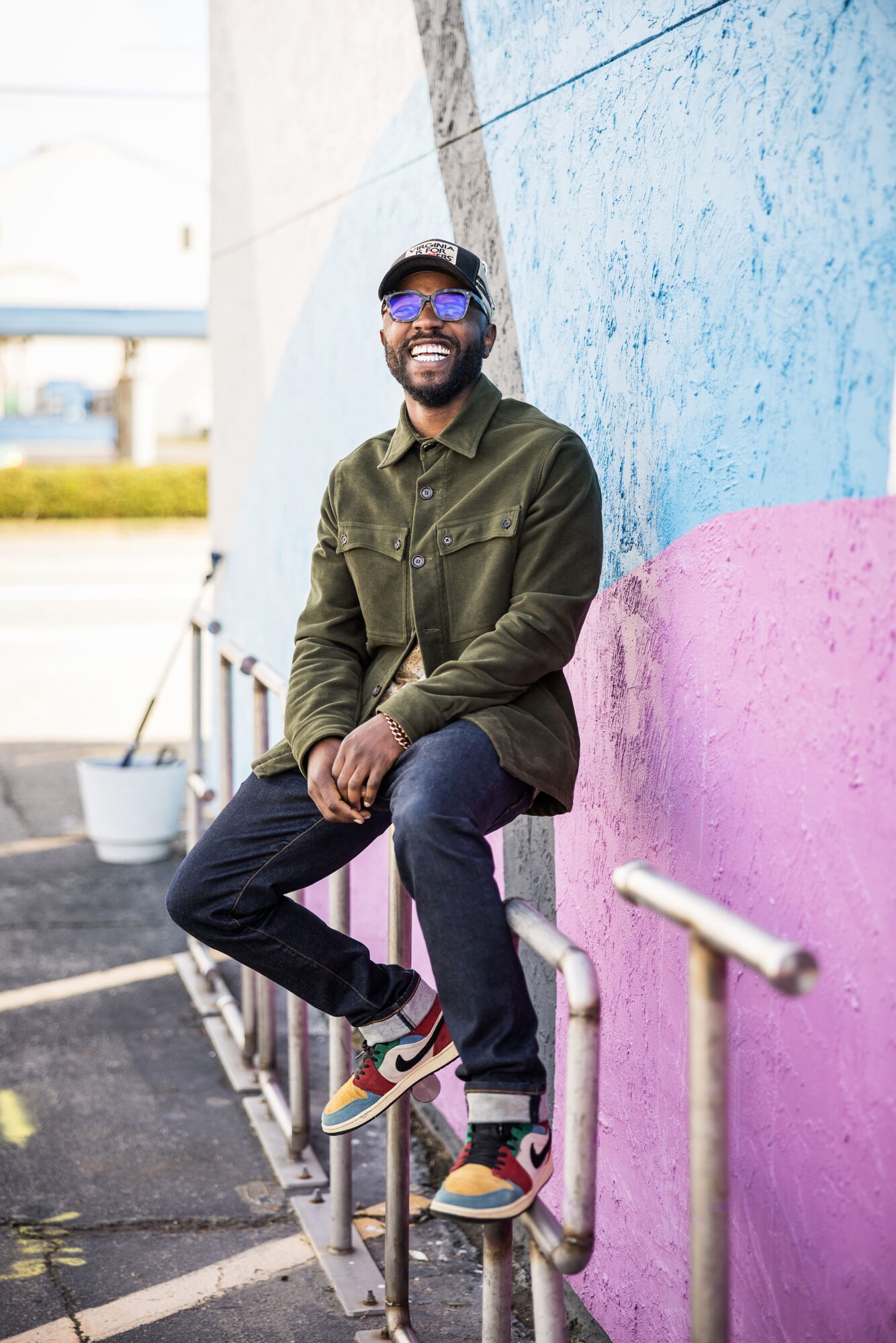 Man with sunglasses and a beard sitting on a railing against a colorful wall, smiling, outdoors.