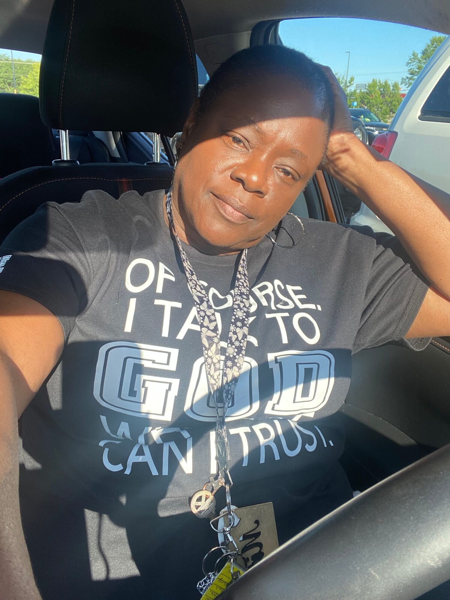 Person sitting in a vehicle, smiling, wearing a black T-shirt with white text, with sunlight and a blue sky visible outside.