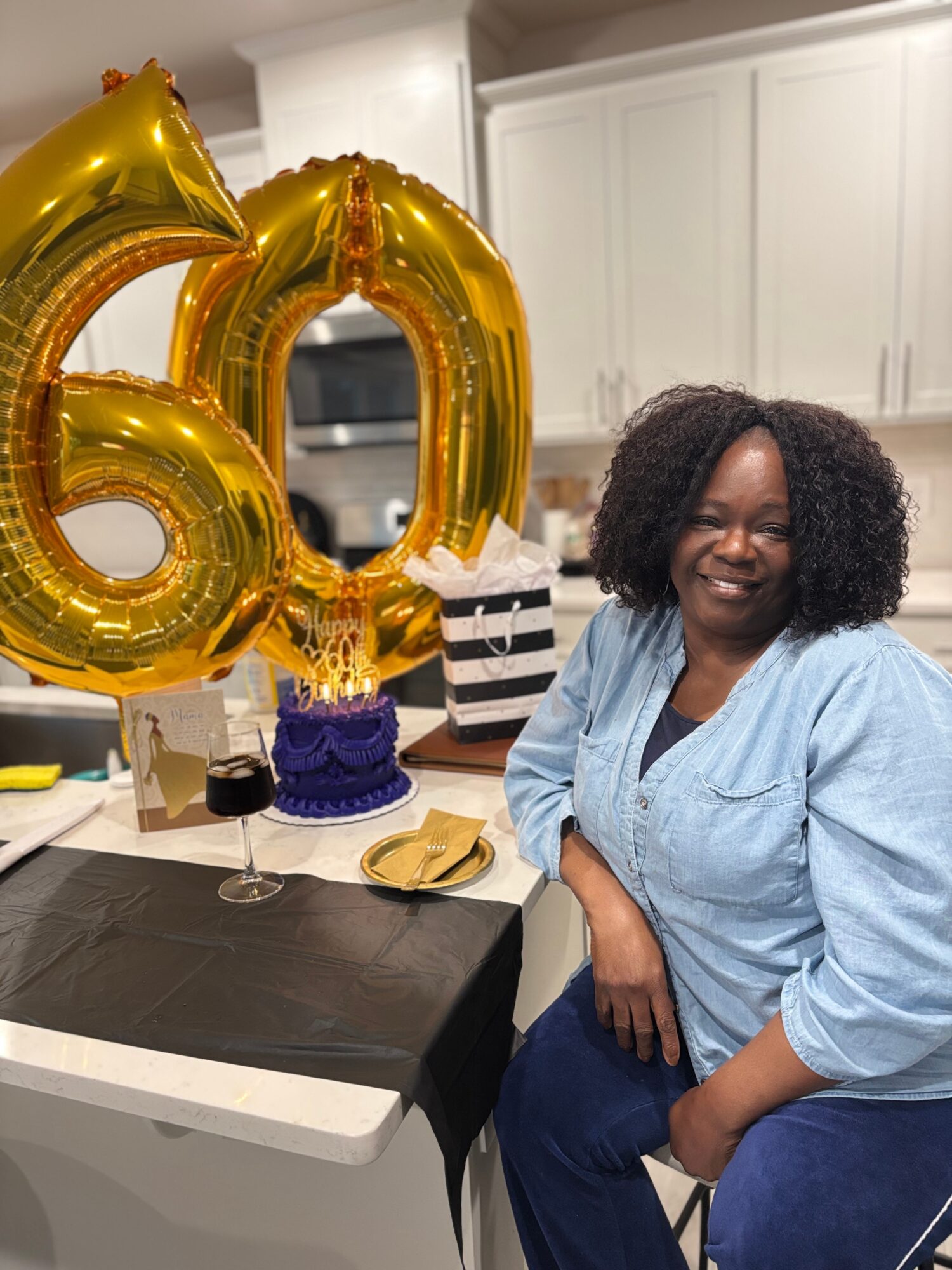Woman smiling at a celebration with large gold '60' balloons in a kitchen, table set with plates and a cake.