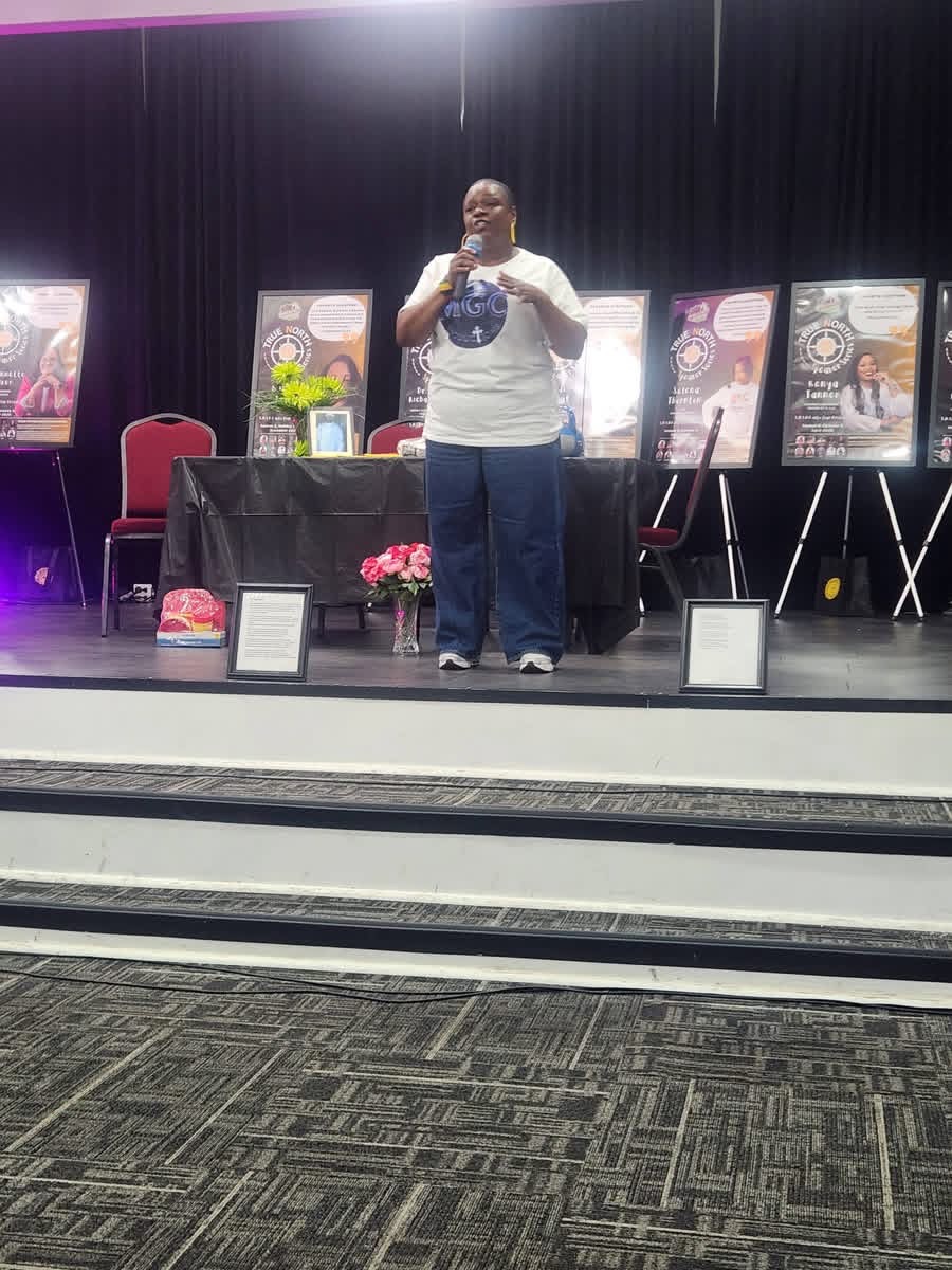 Person standing on stage with books displayed behind, holding a microphone, in a conference or event setting.