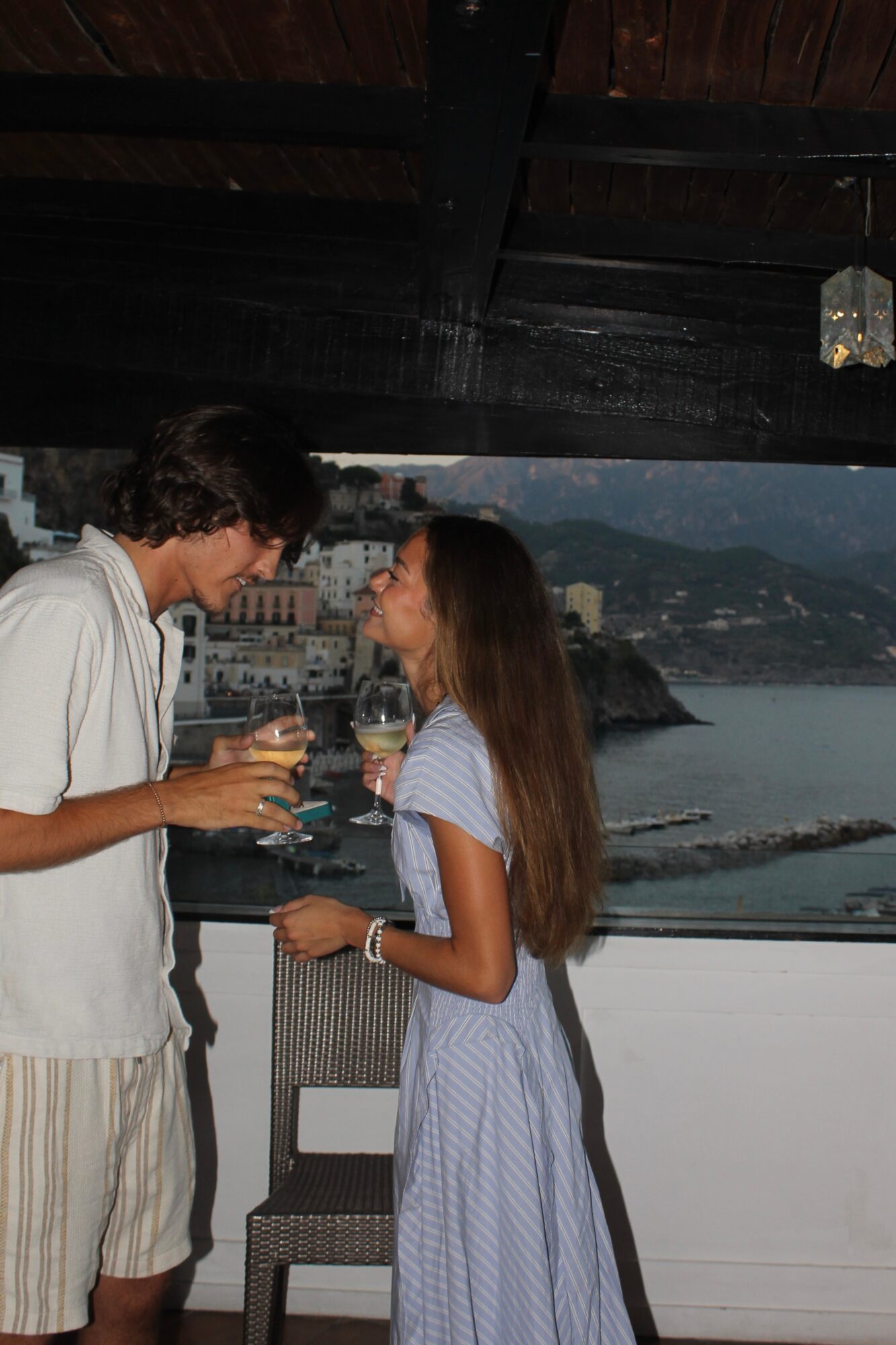 Two people holding glasses and talking indoors with a view of water and buildings outside window.