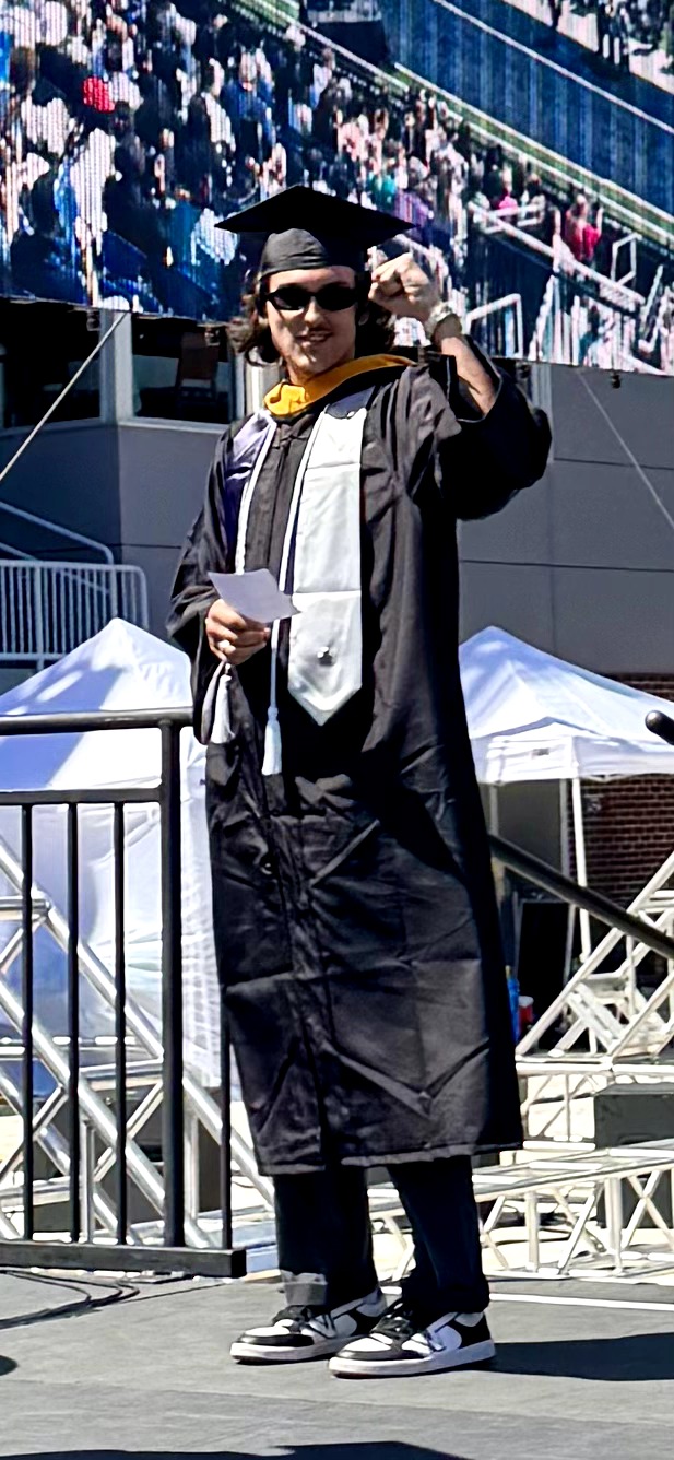 Person in graduation gown and cap holding a diploma, standing outdoors with a crowd in the background.