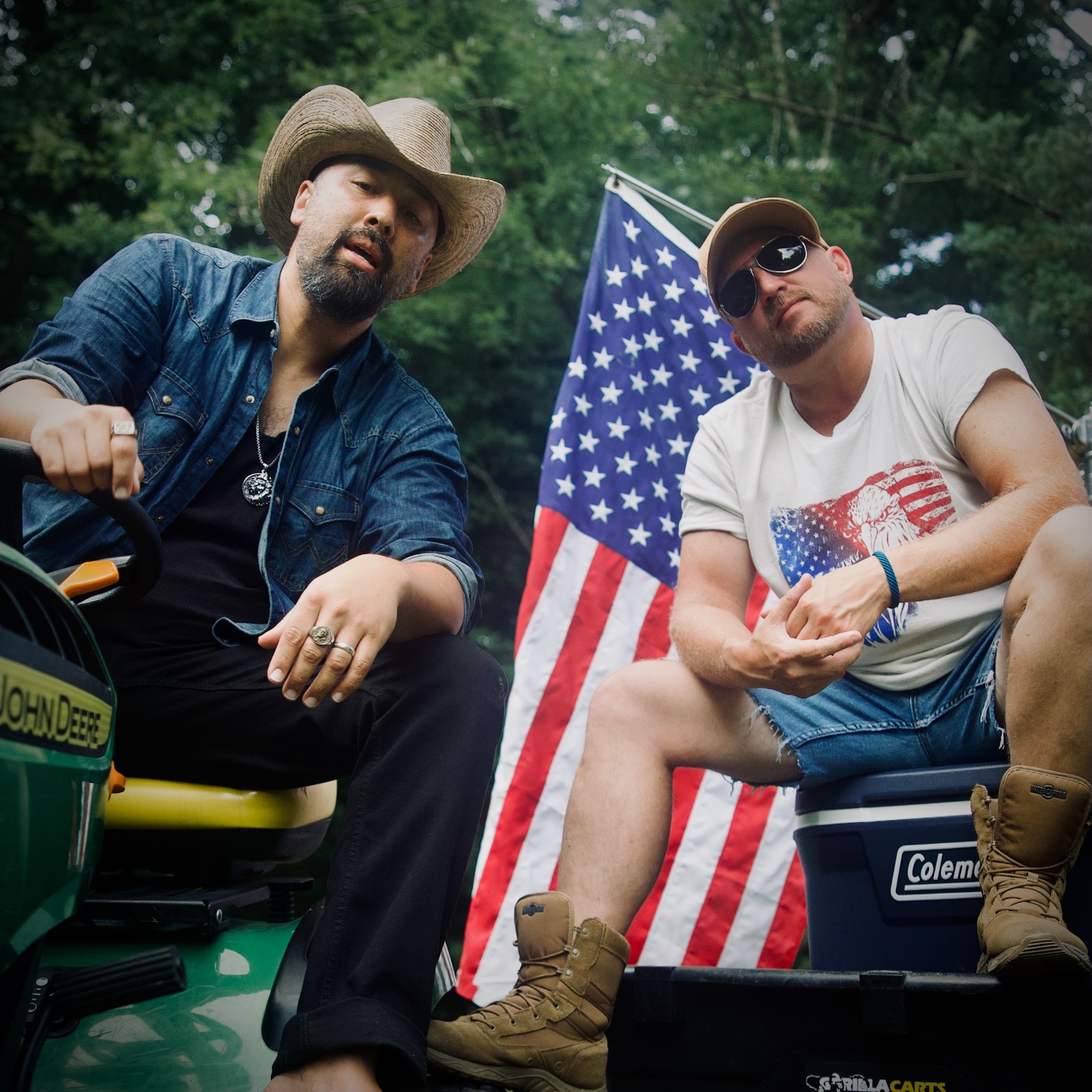 Two men sit outdoors with an American flag, one wearing a cowboy hat and sunglasses, the other in a baseball cap and sunglasses.