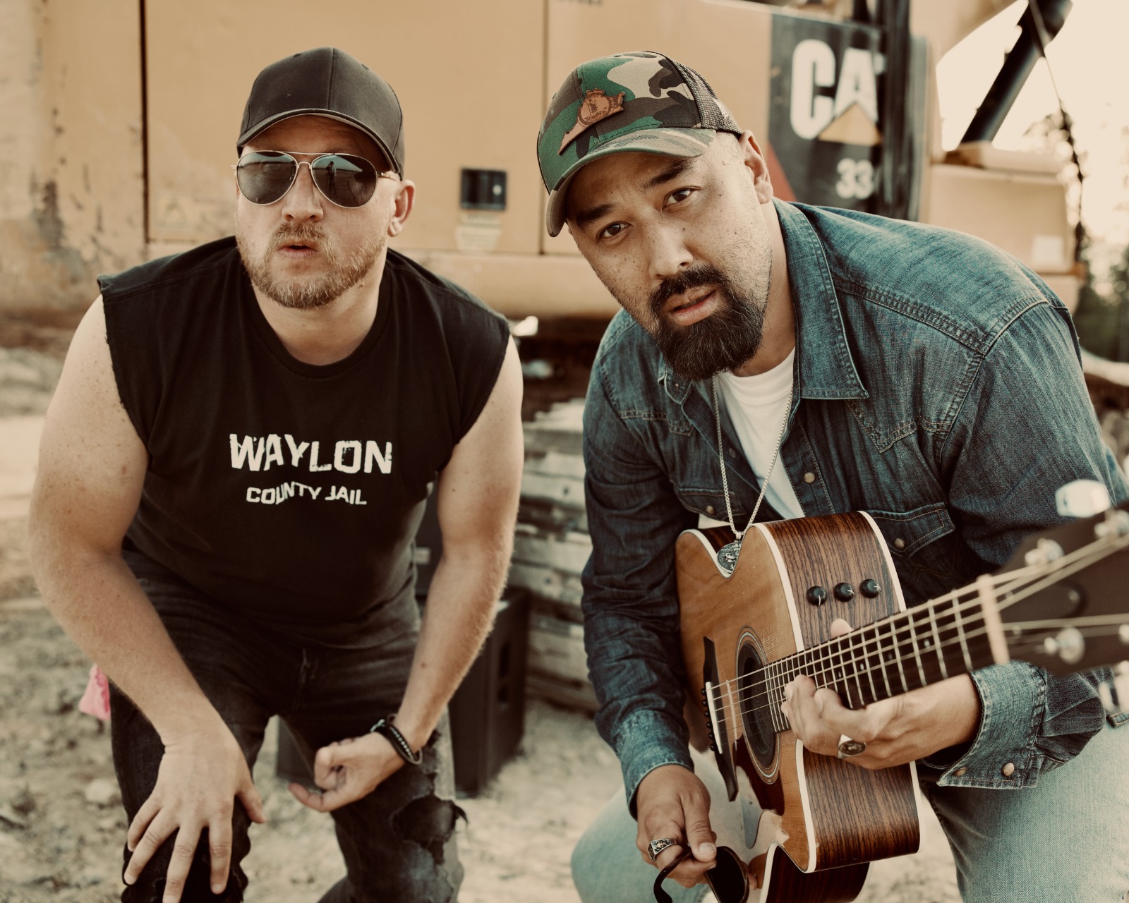 Two men, one wearing sunglasses and a cap, the other with a beard and a cap, holding a guitar outdoors.