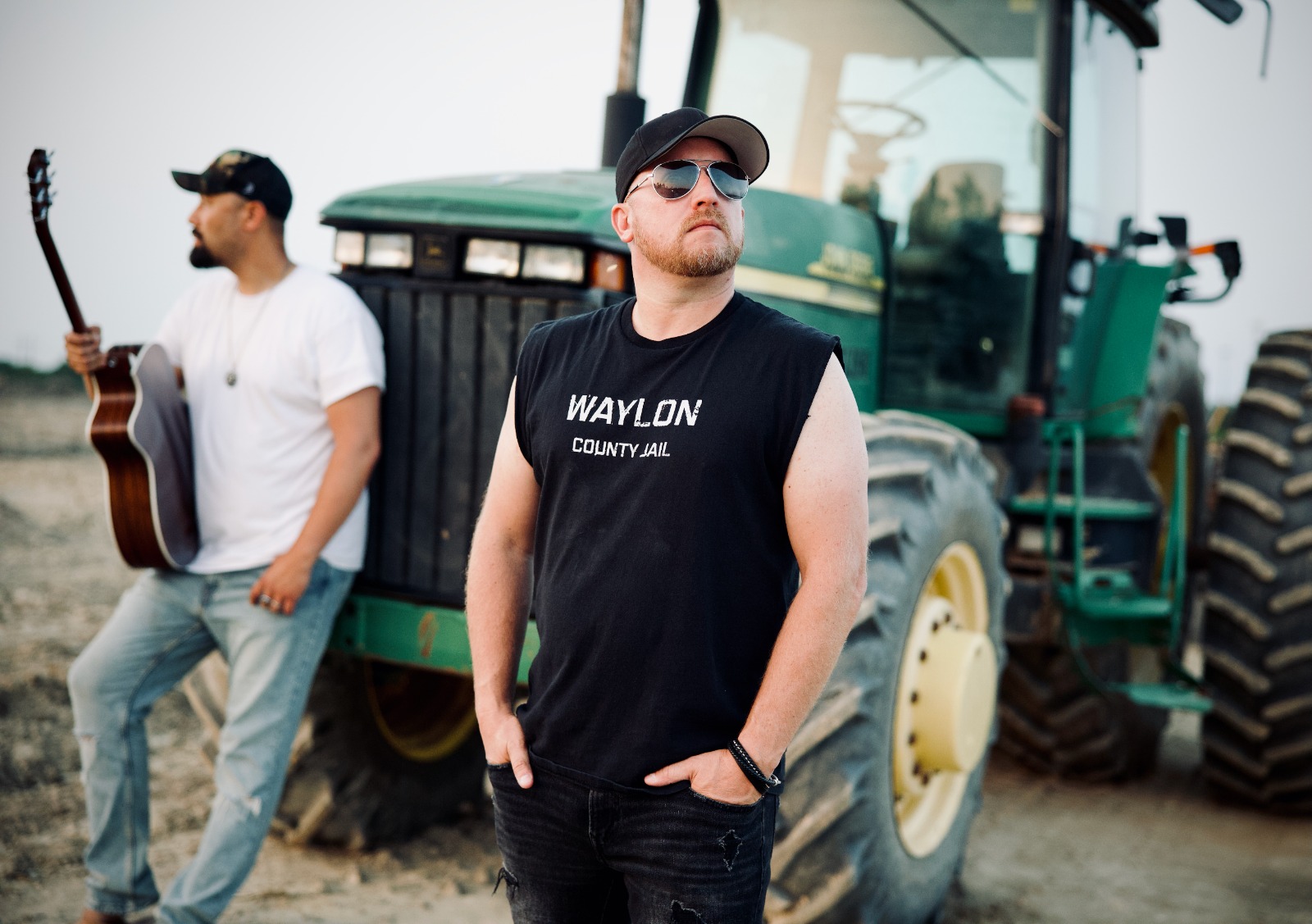 Two men stand outdoors near a large green tractor, one wearing sunglasses and a sleeveless shirt, the other with a guitar.