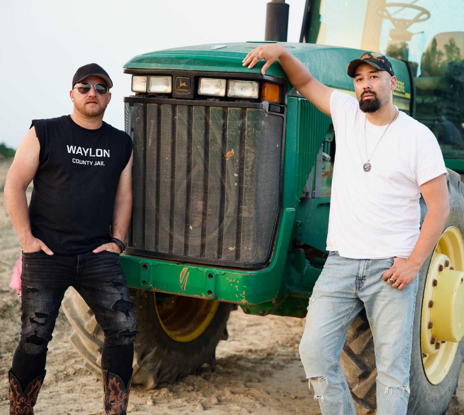 Two men stand beside a green tractor outdoors, one resting arm on the tractor's hood, both looking at the camera.