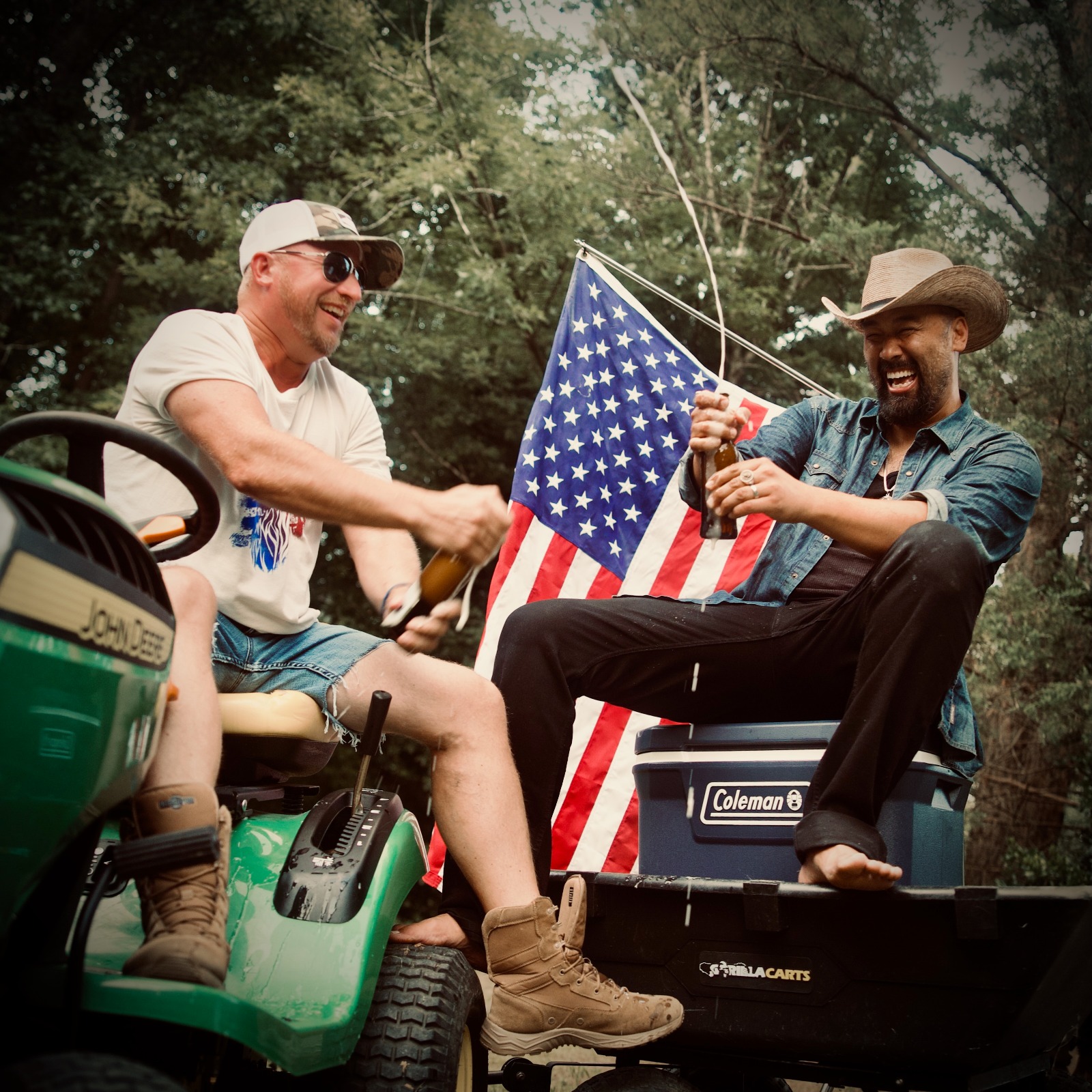Two men smiling and shaking hands outdoors, one holding an American flag, sitting on a cooler and a golf cart, trees in background.