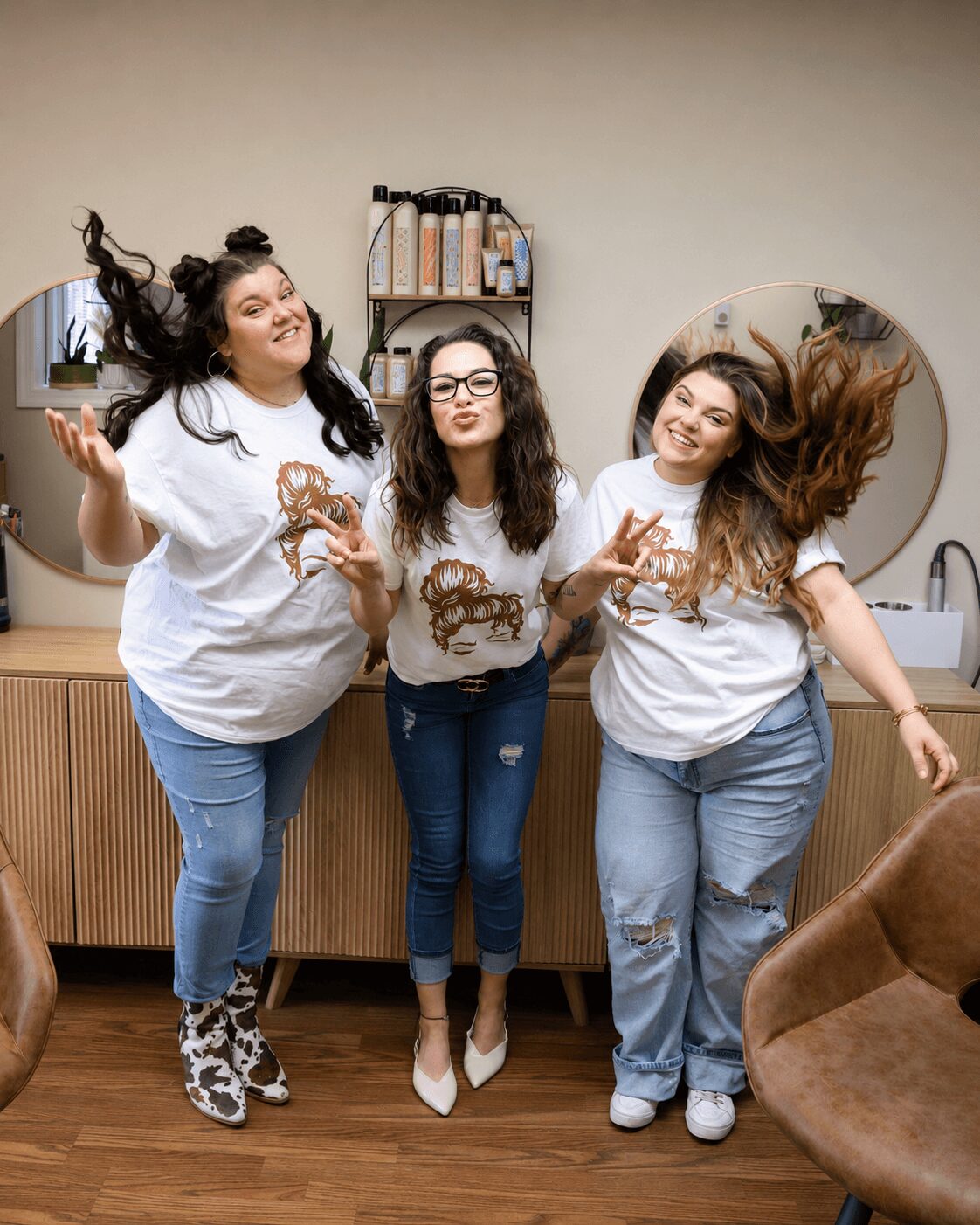 Three women with long hair, wearing white shirts and jeans, pose and smile in a room with mirrors and a wooden cabinet.