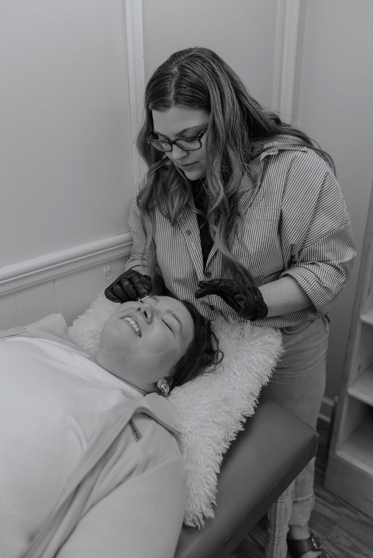 A woman with glasses and gloves leans over a person lying on a bed, smiling and looking at them.