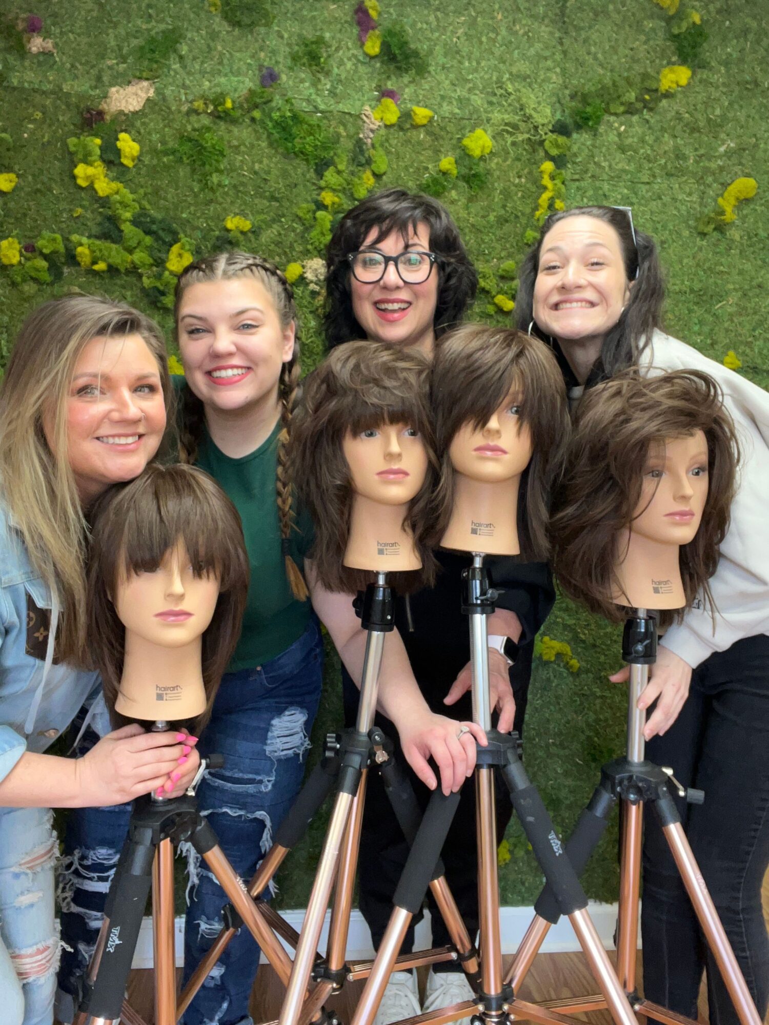 Group of six women outdoors with yellow flowers and green grass, posing for a photo, some with hands on camera equipment.