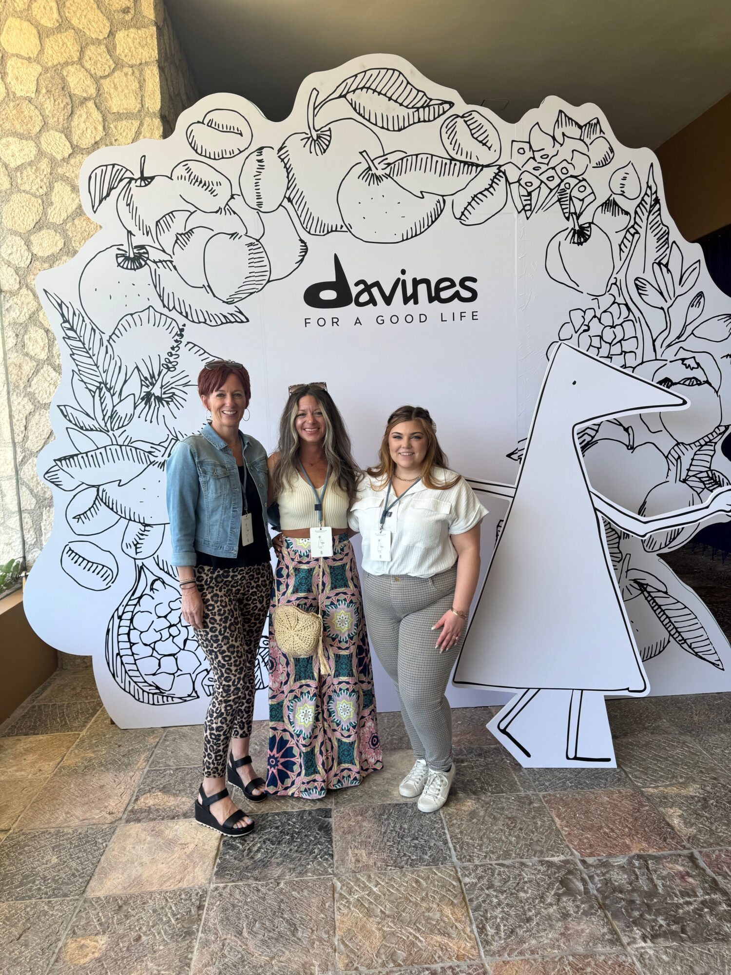 Three women stand in front of a large, decorated cutout with the word 'Davines' and floral illustrations, indoors.